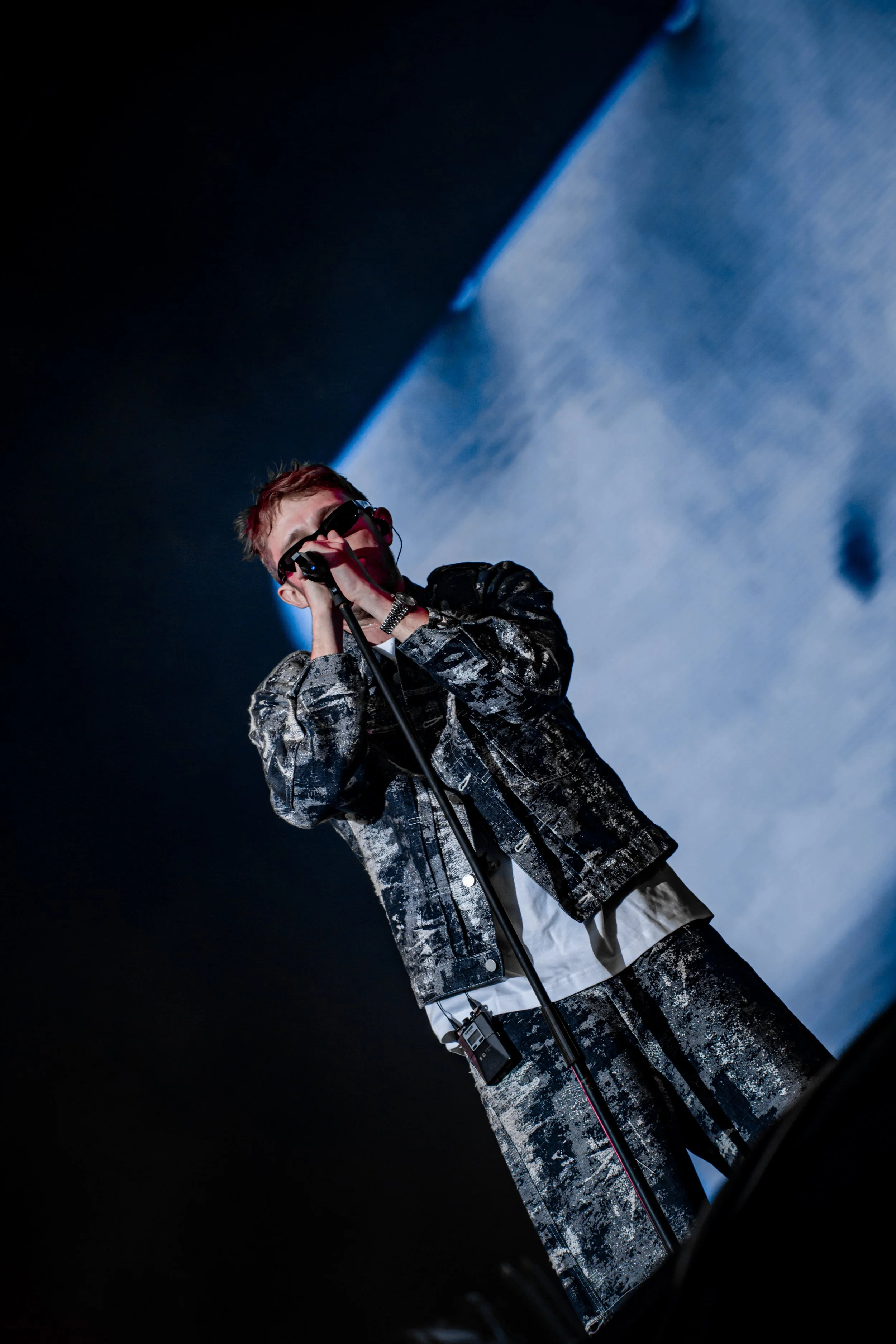 Un jeune homme portant des lunettes noires chantant dans un micro lors d'une performance sur scène, vêtu d'une veste à motifs camouflage noir et blanc.