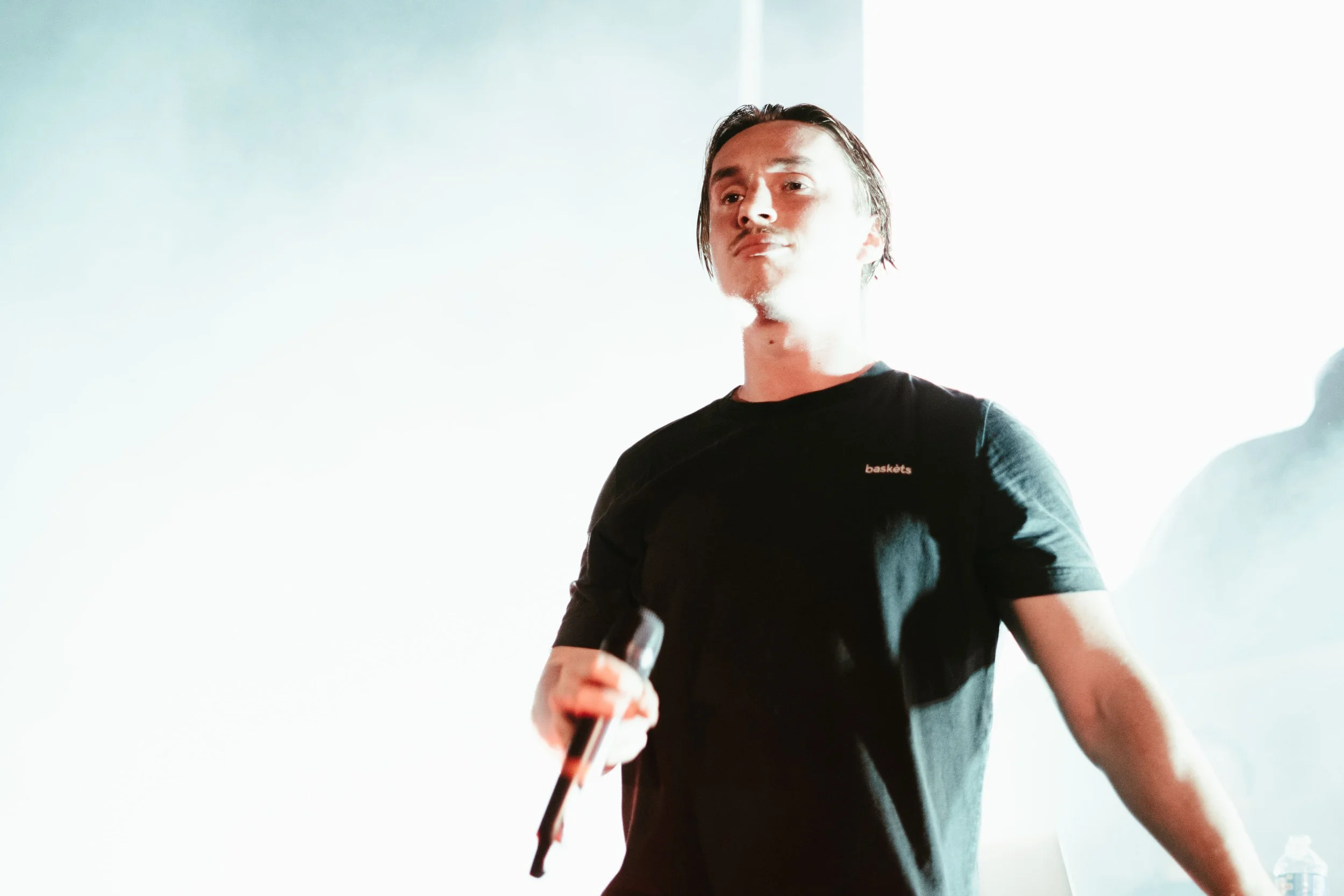 Un homme avec des cheveux longs porte un t-shirt noir et tient un microphone, se tenant dans un environnement lumineux.