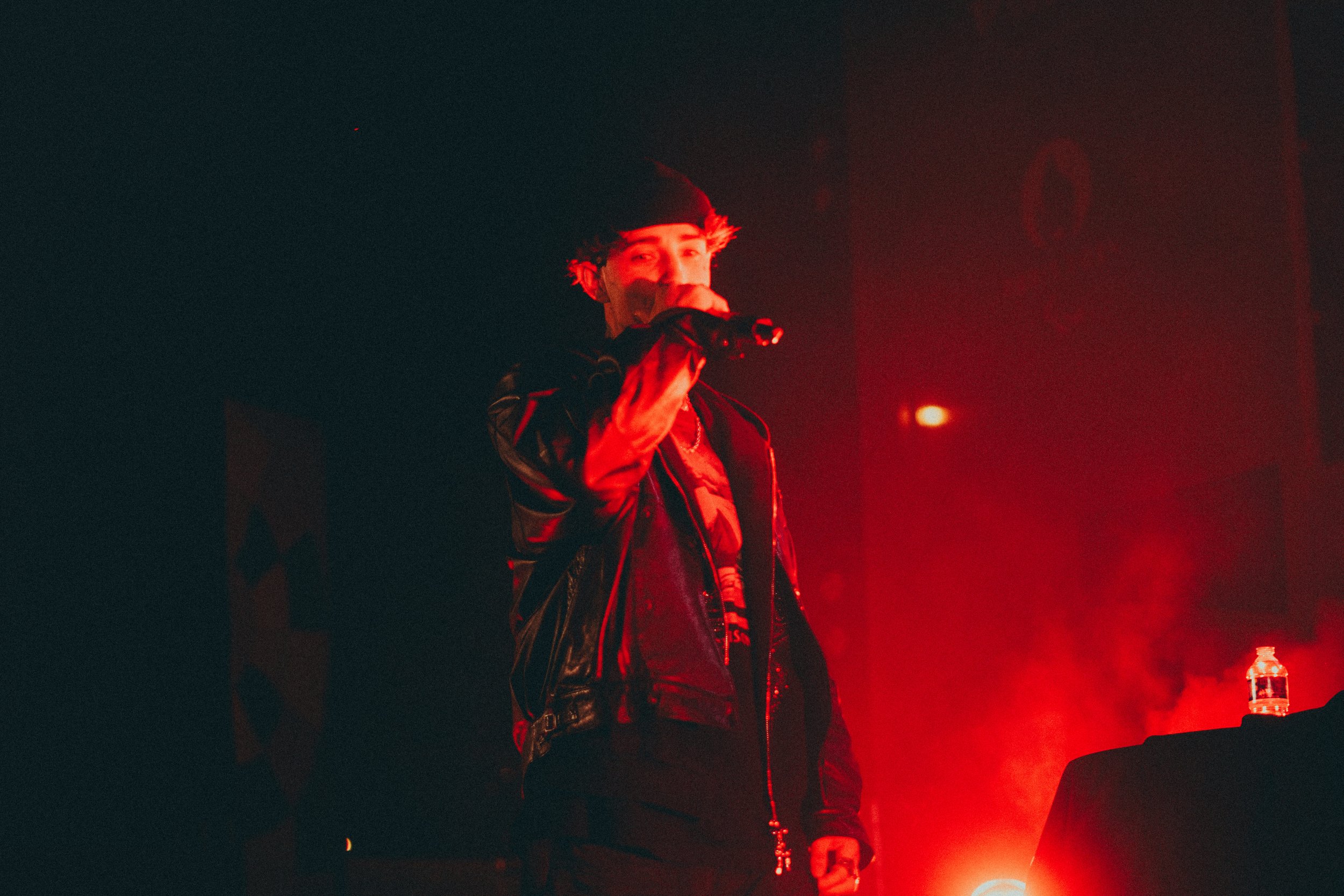 Un jeune homme portant un chapeau noir et une veste en cuir noir, tenant un micro lors d'une performance sur scène, entouré d'une lumière rouge