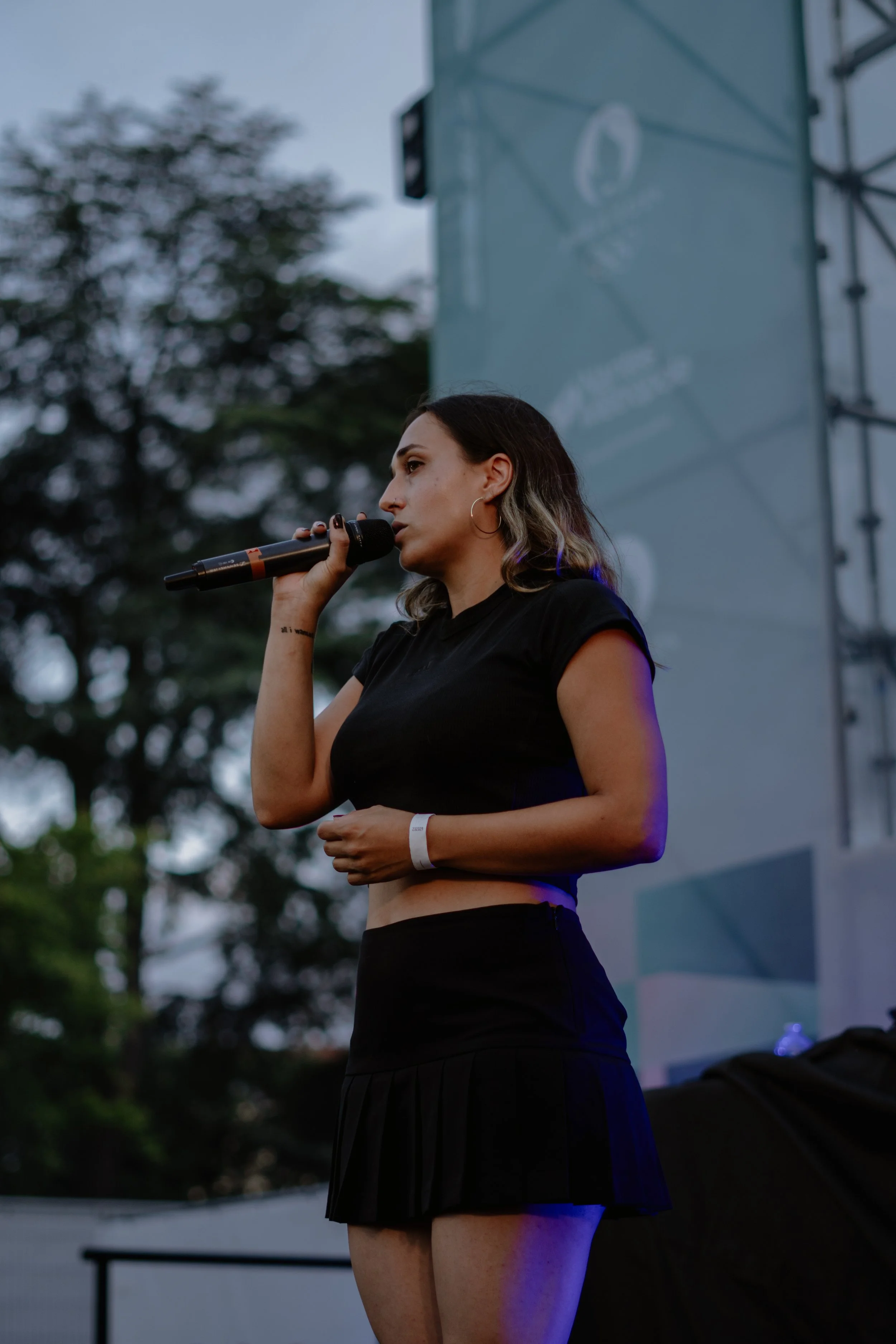 Une femme parle dans un microphone lors d'un événement en plein air, avec un arrière-plan d'arbres et un écran géant.