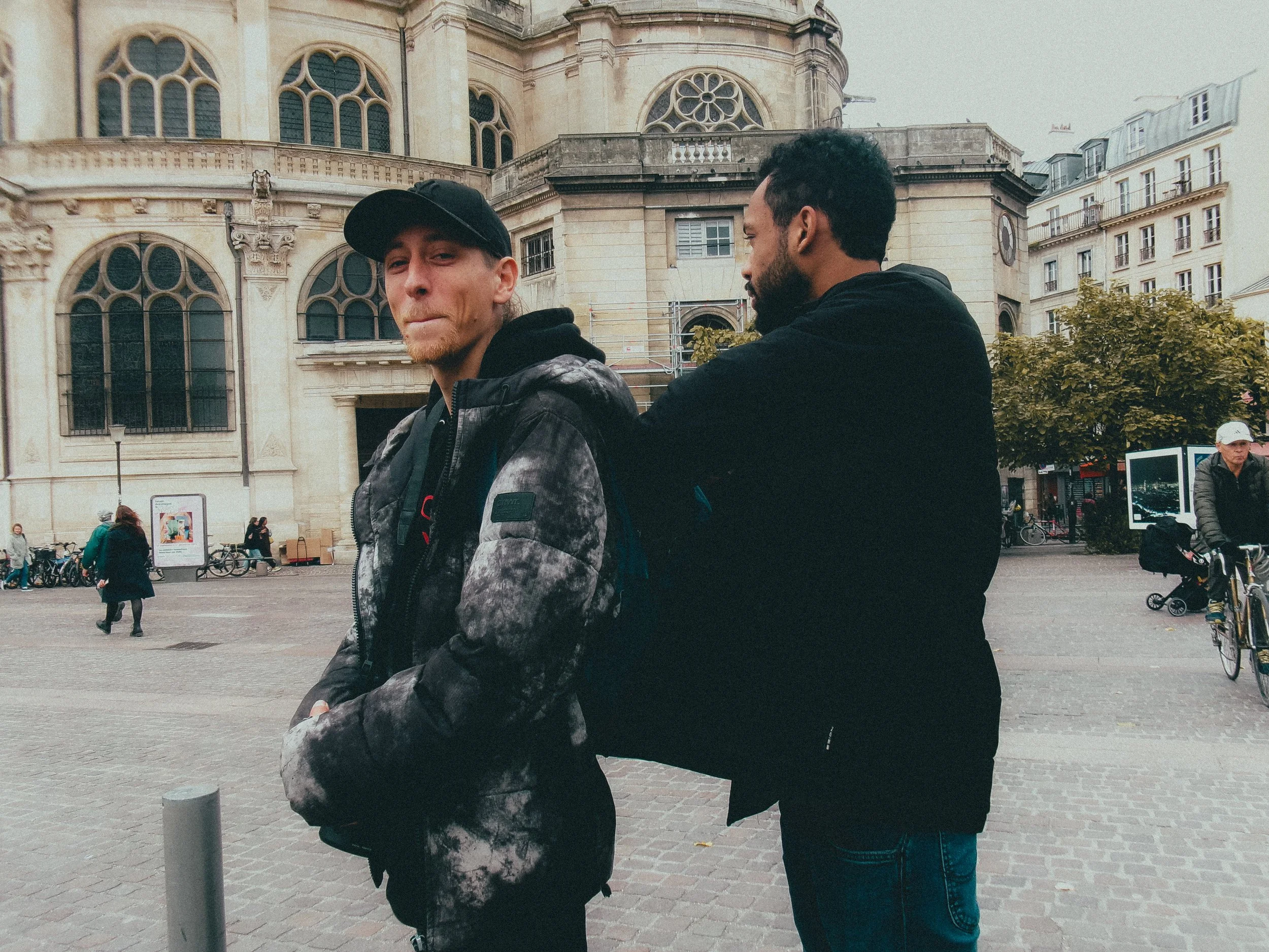 Deux jeunes hommes debout dans une place urbaine, l'un regarde la caméra avec un léger sourire, l'autre regarde vers l'avant. En arrière-plan, un bâtiment historique avec des fenêtres arched. Il y a aussi des vélos, des piétons et un arbre.