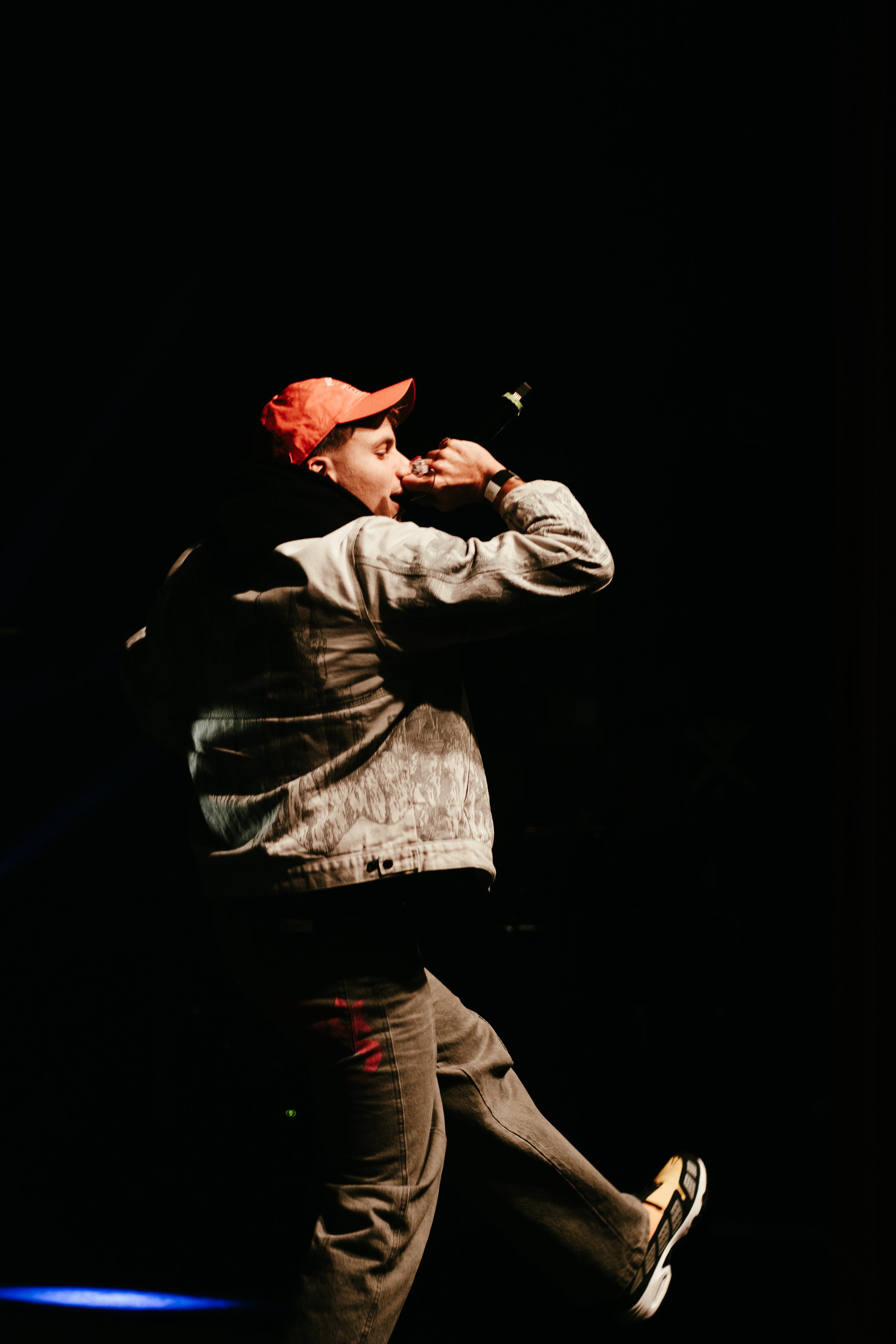 Un jeune homme portant une casquette rouge, un sweat à capuche et un pantalon cargo, en train de boire lors d'une performance sur scène, avec un fond noir.
