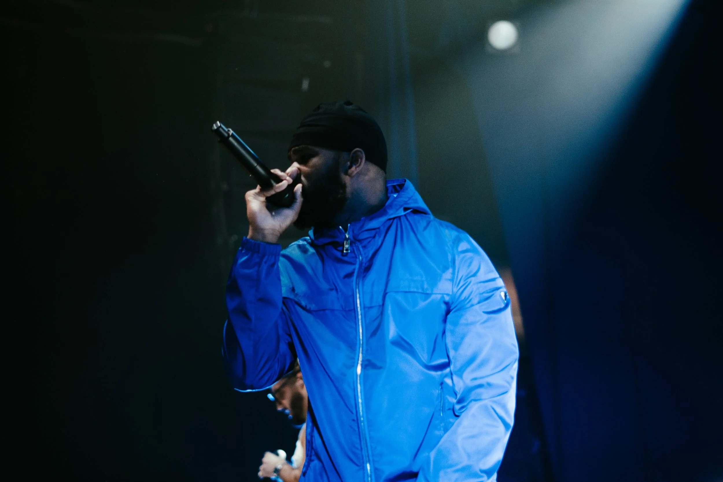 Un homme portant un bonnet noir et une veste bleue tient un micro près de sa bouche dans une pièce sombre avec une lumière dirigée vers lui.