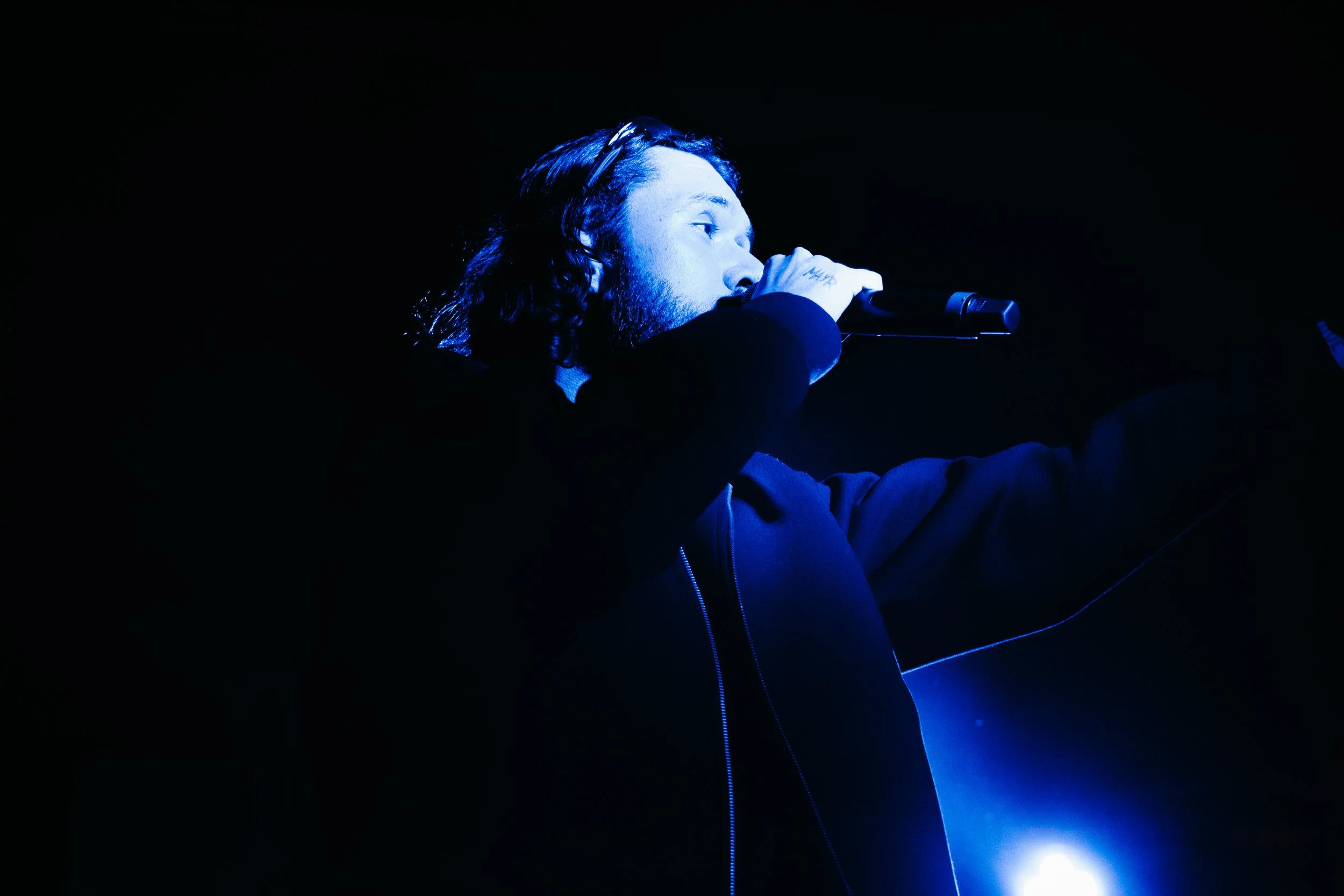 Un homme avec des cheveux longs et une barbe, chantant ou parlant dans un microphone dans une scène sombre, éclairée par une lumière bleue.