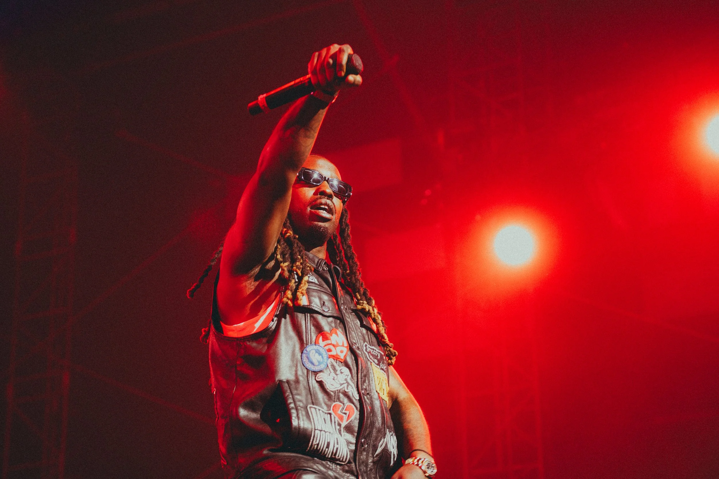Un homme avec dreadlocks portant des lunettes de soleil, une veste en cuir ornée de badges, tenant un microphone, sur scène sous un éclairage rouge.