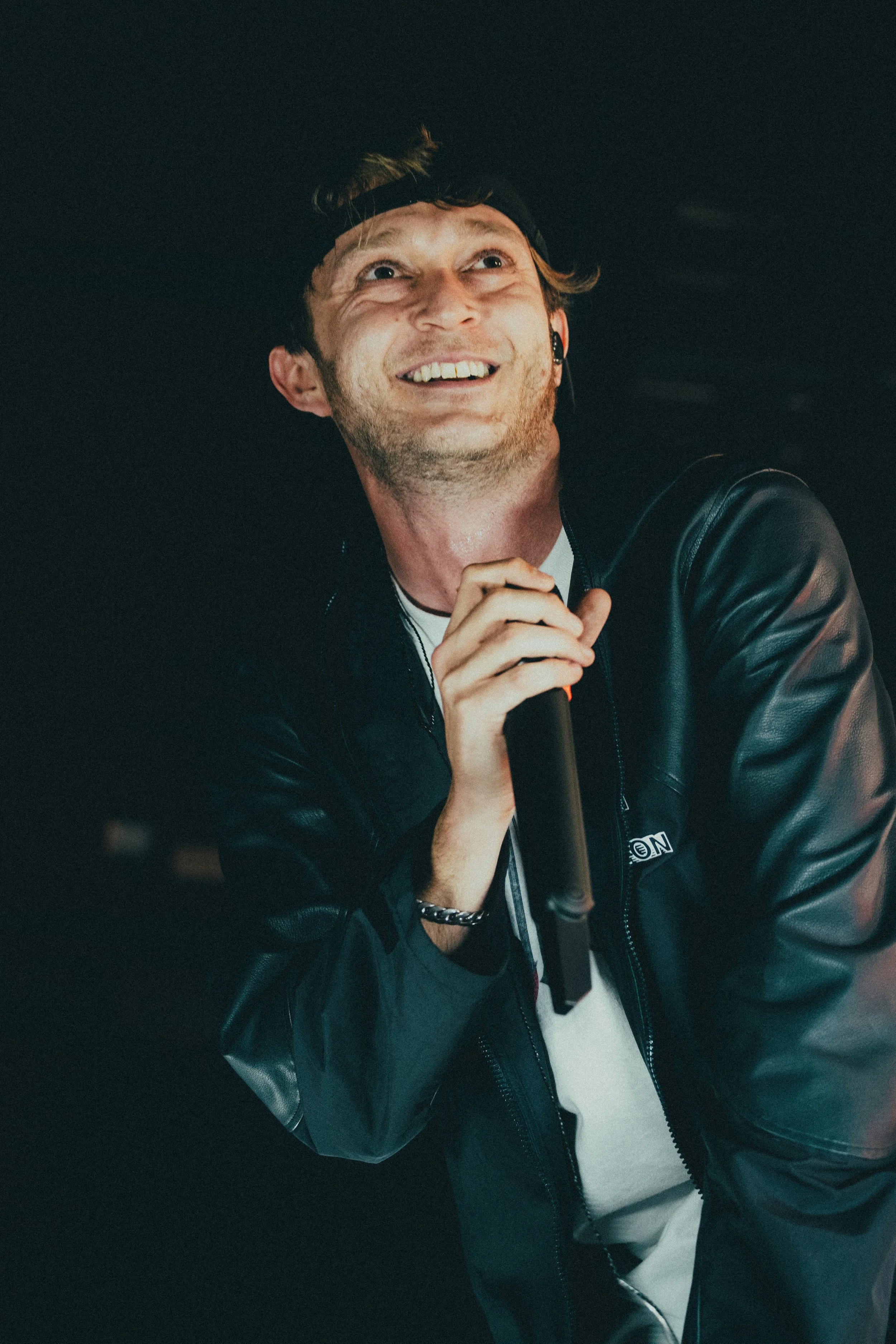 Jeune homme chantant ou parlant en public avec un micro, portant une veste en cuir, souriant et portant un bandeau ou une casquette