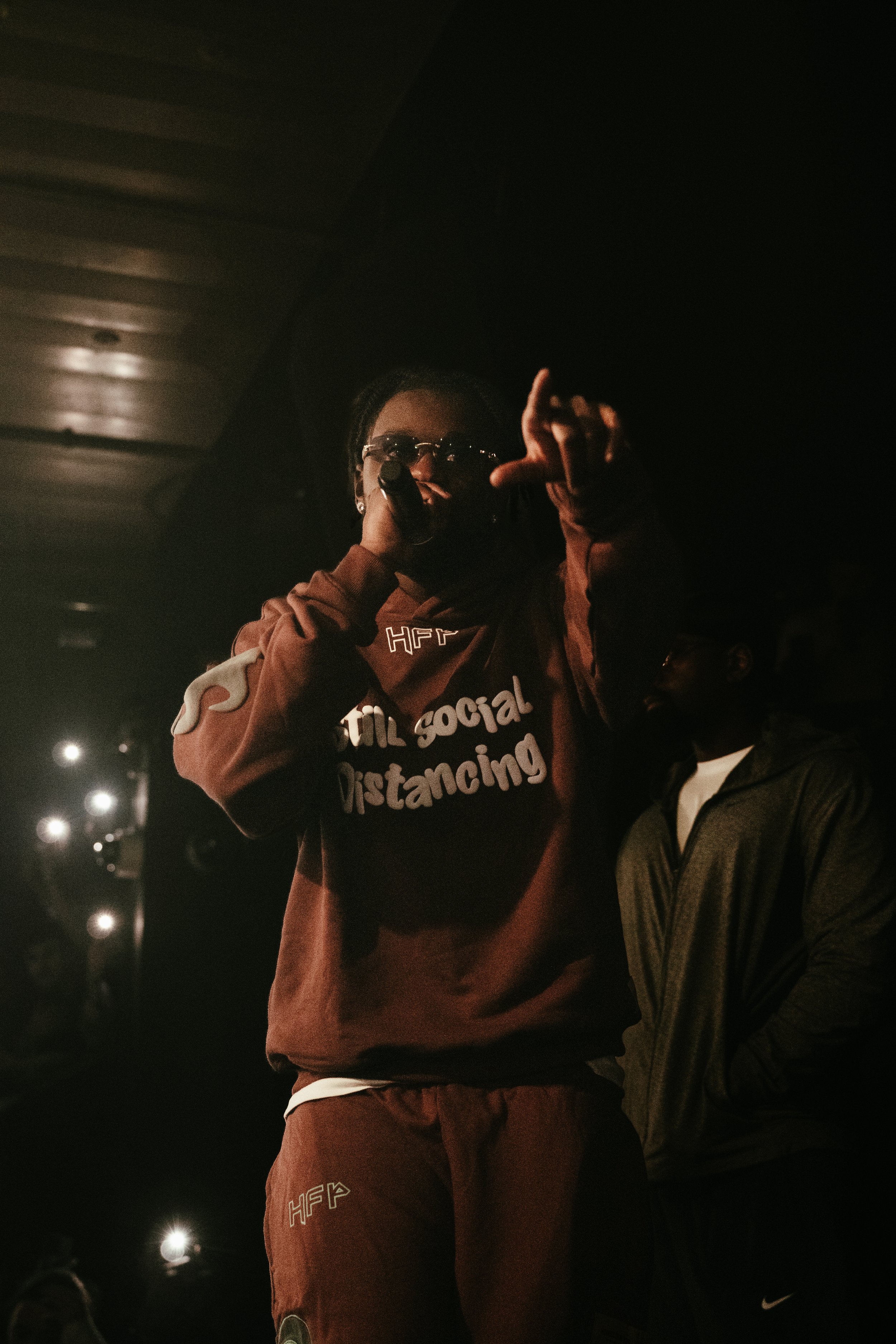 Une personne portant un sweat marron avec l'inscription 'Still social distancing' parle dans un micro, en face d'un public dans une ambiance sombre.