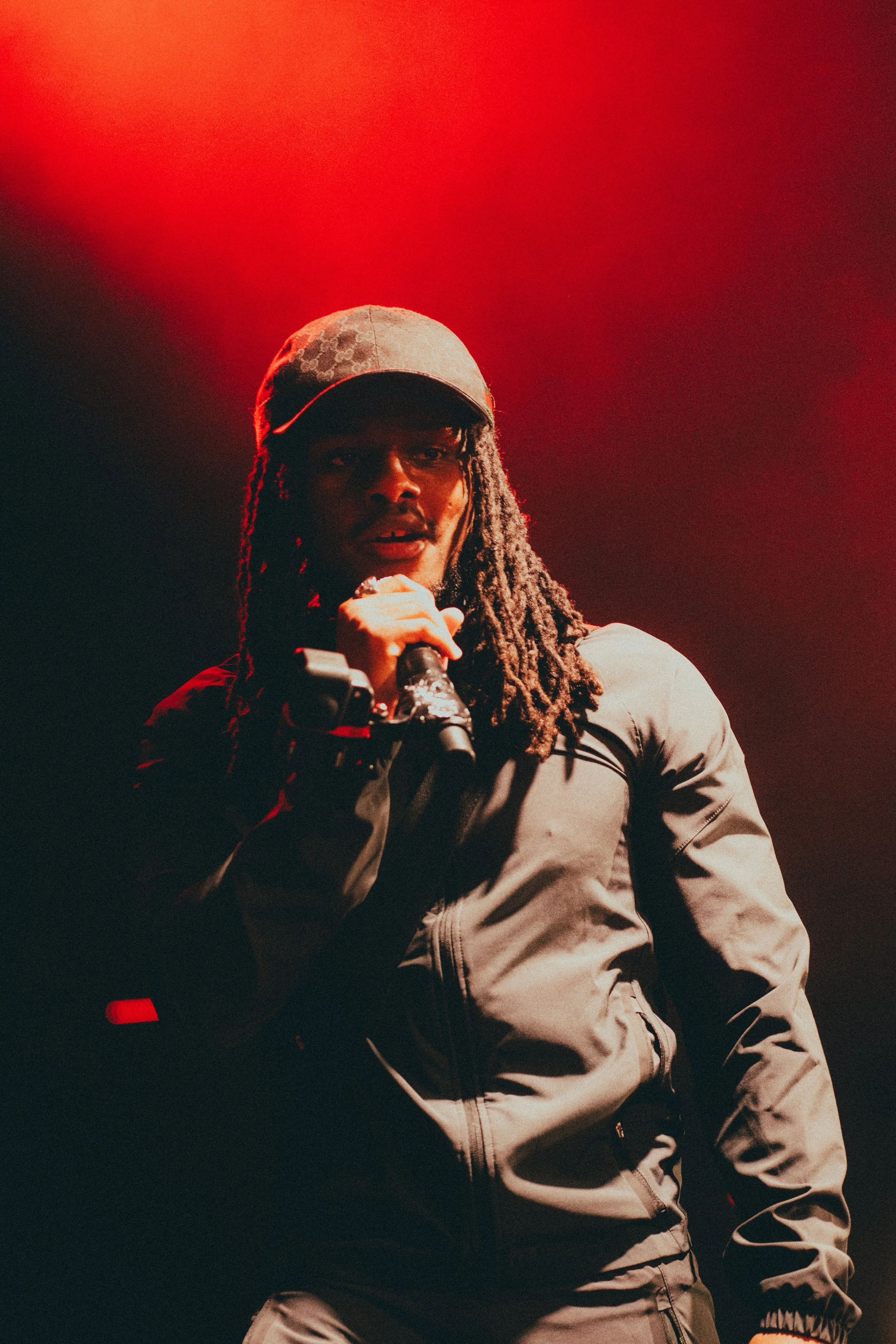 Un homme avec des dreadlocks porte une casquette et une veste foncée, tenant un microphone lors d'une performance sur scène, éclairé par des lumières rouges.