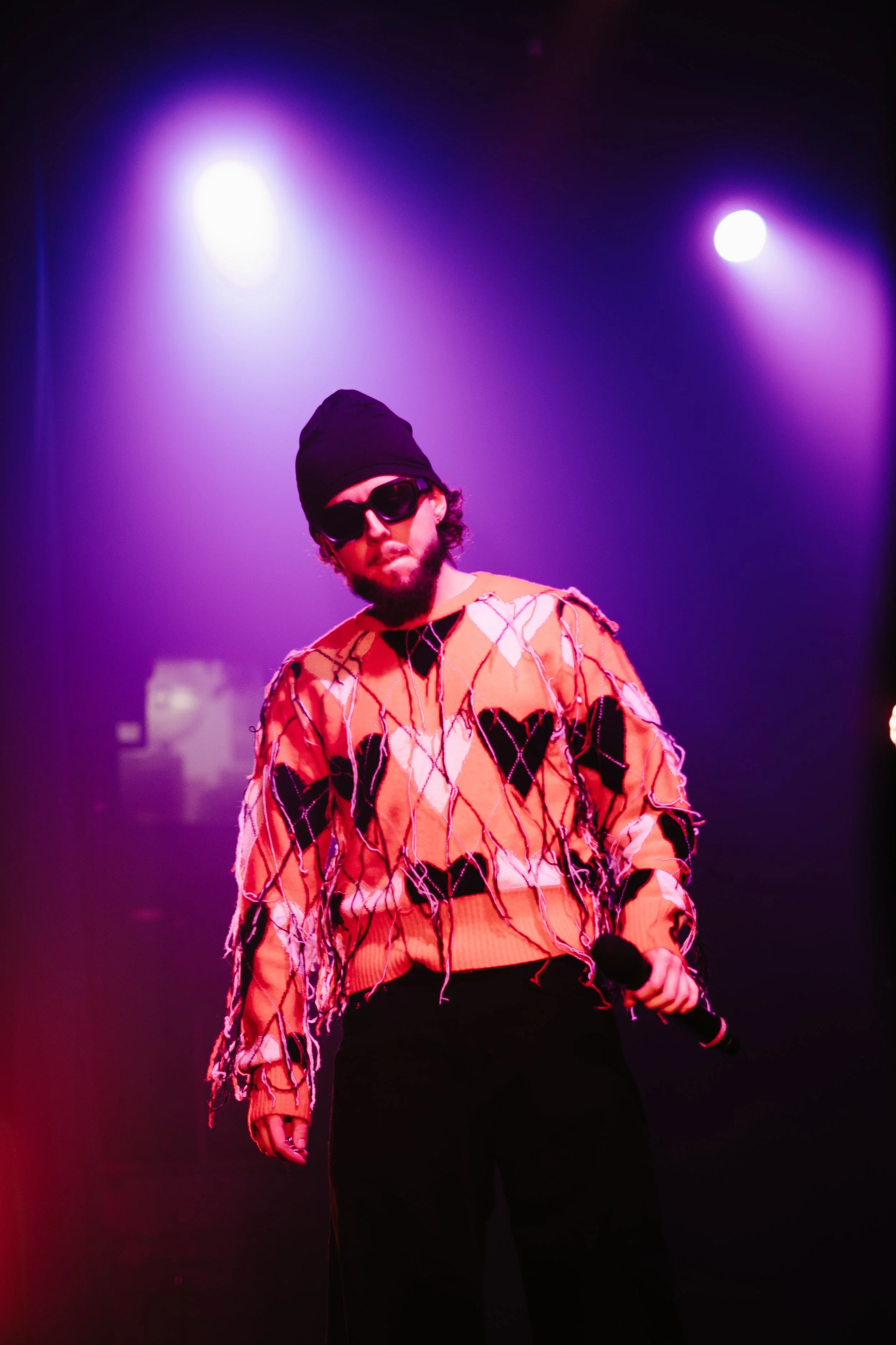 Un homme avec des lunettes de soleil, un bonnet noir, une barbe, tenant un micro, sous un éclairage violet sur scène.