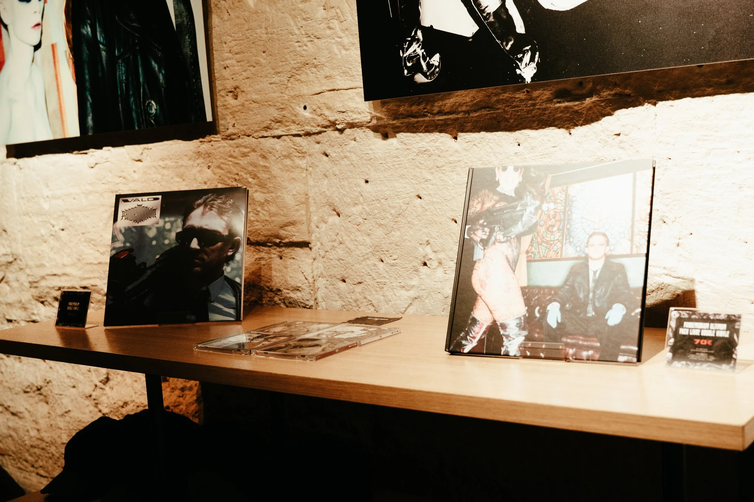 Affiches et photographies d'art exposées sur un mur en pierre dans une galerie, avec des personnages en costumes et une ambiance artistique.