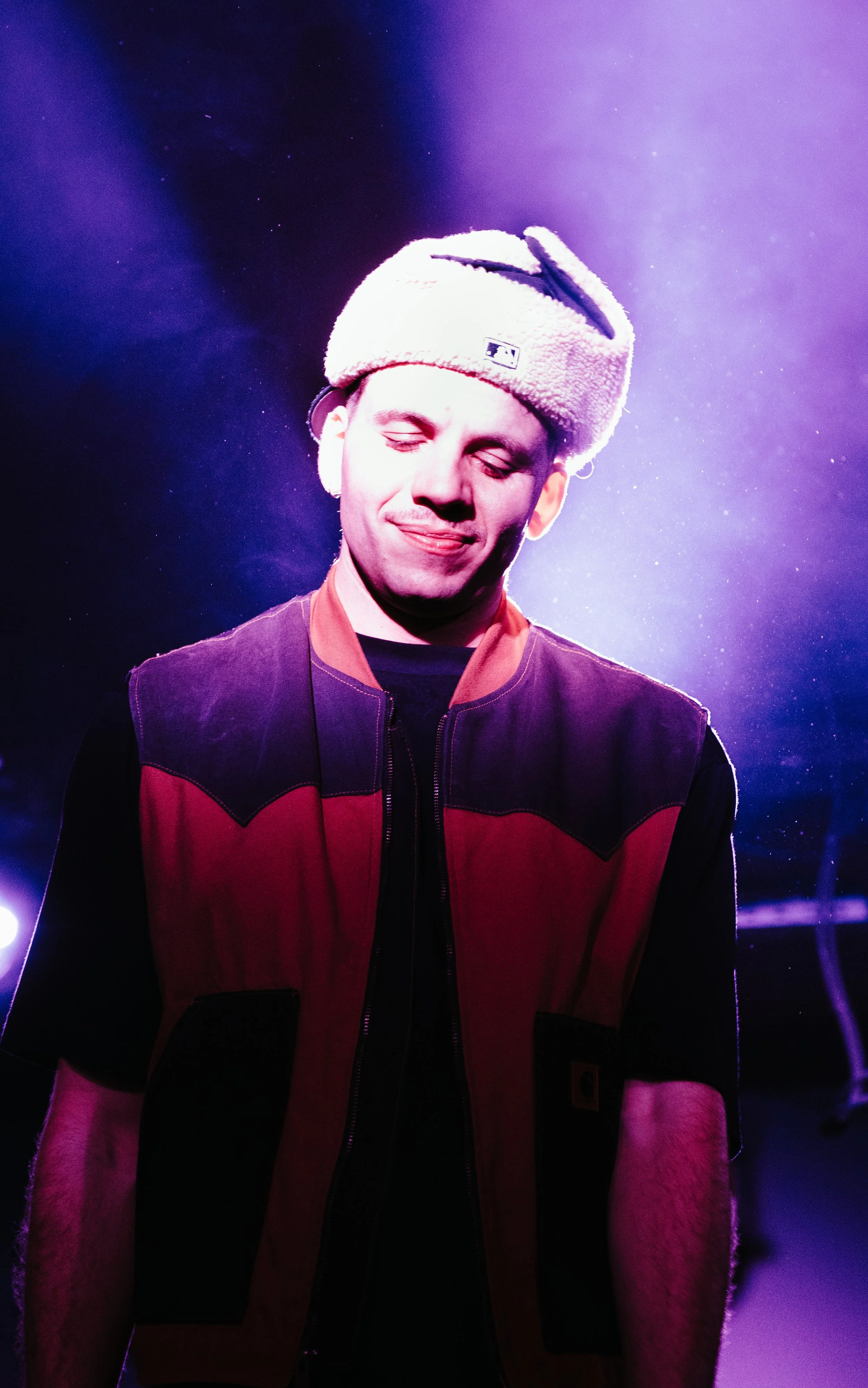 Un homme souriant portant une veste noire et rouge, ainsi qu'un bonnet en peluche beige qui ressemble à une oreille d'animal, avec un fond de lumières violettes et bleues.