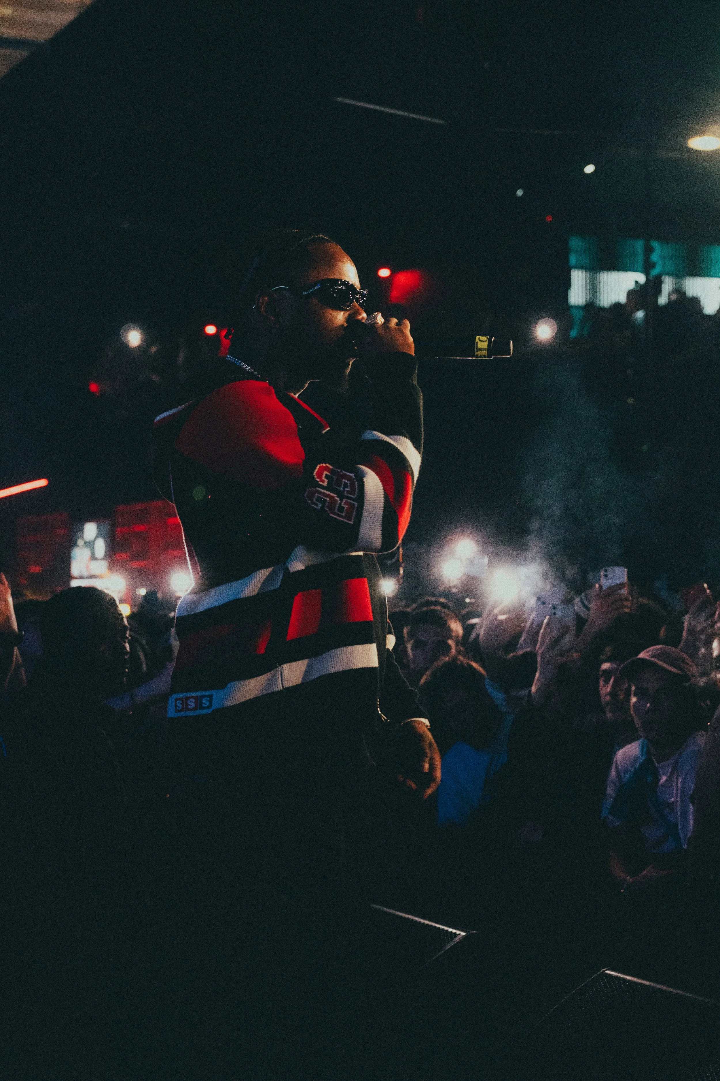 Un artiste en train de performer avec un micro dans une ambiance sombre, entouré par un public assis dans une senteur de concert ou de scène artistique.