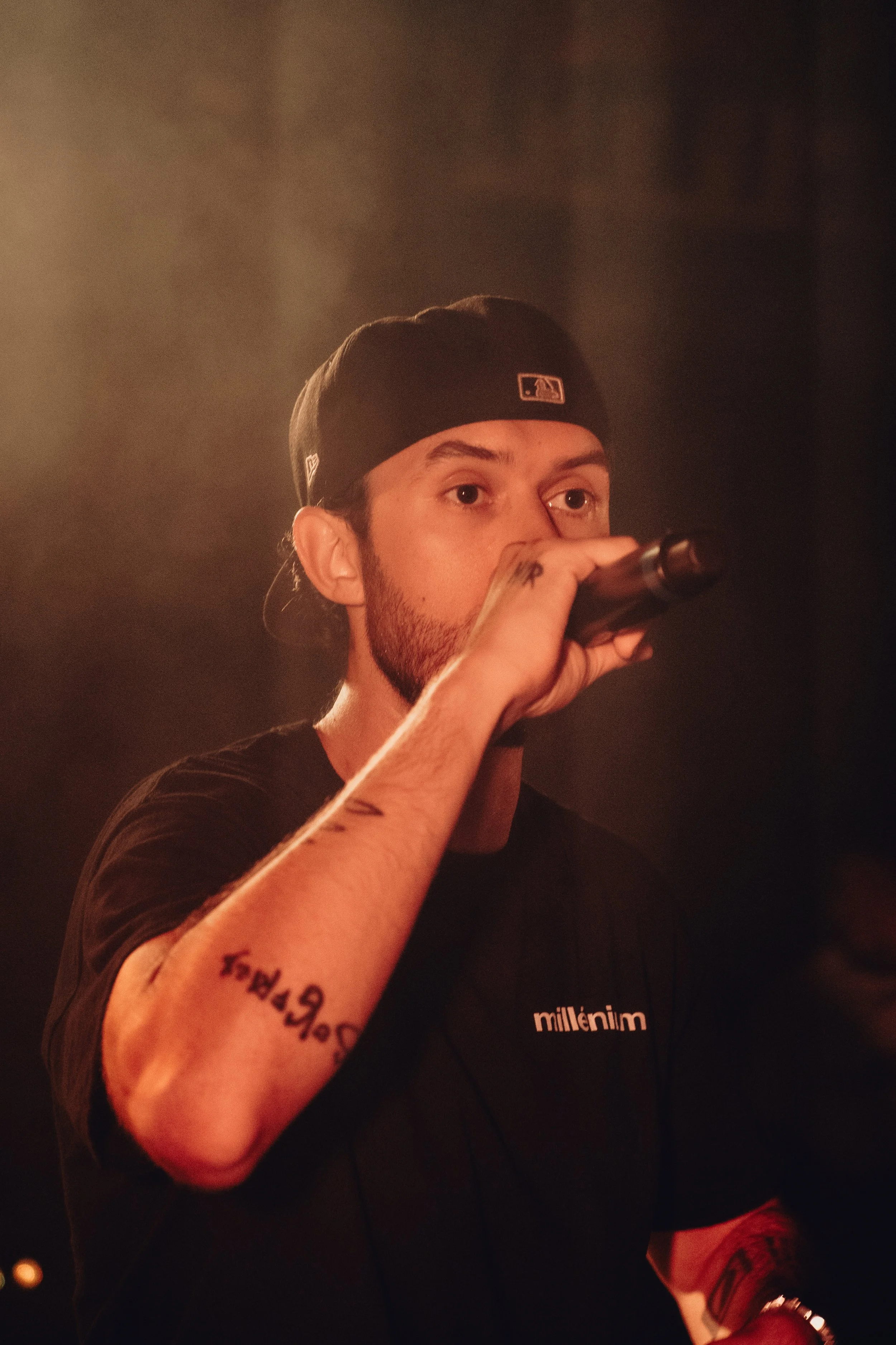 Un homme portant une casquette noire, tenant un microphone, portant un t-shirt noir avec l'inscription 'millènim', avec des tatouages visibles sur le bras.