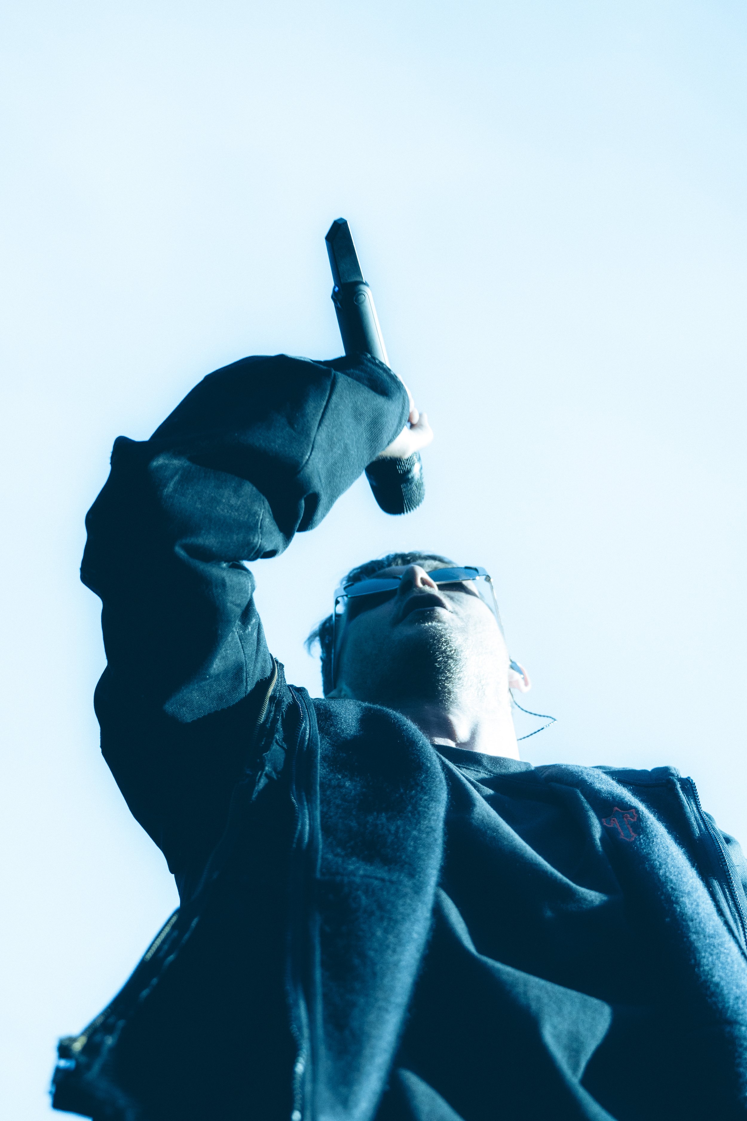 Une personne portant des lunettes, tenant un microphone, vue de dessous contre un ciel clair.