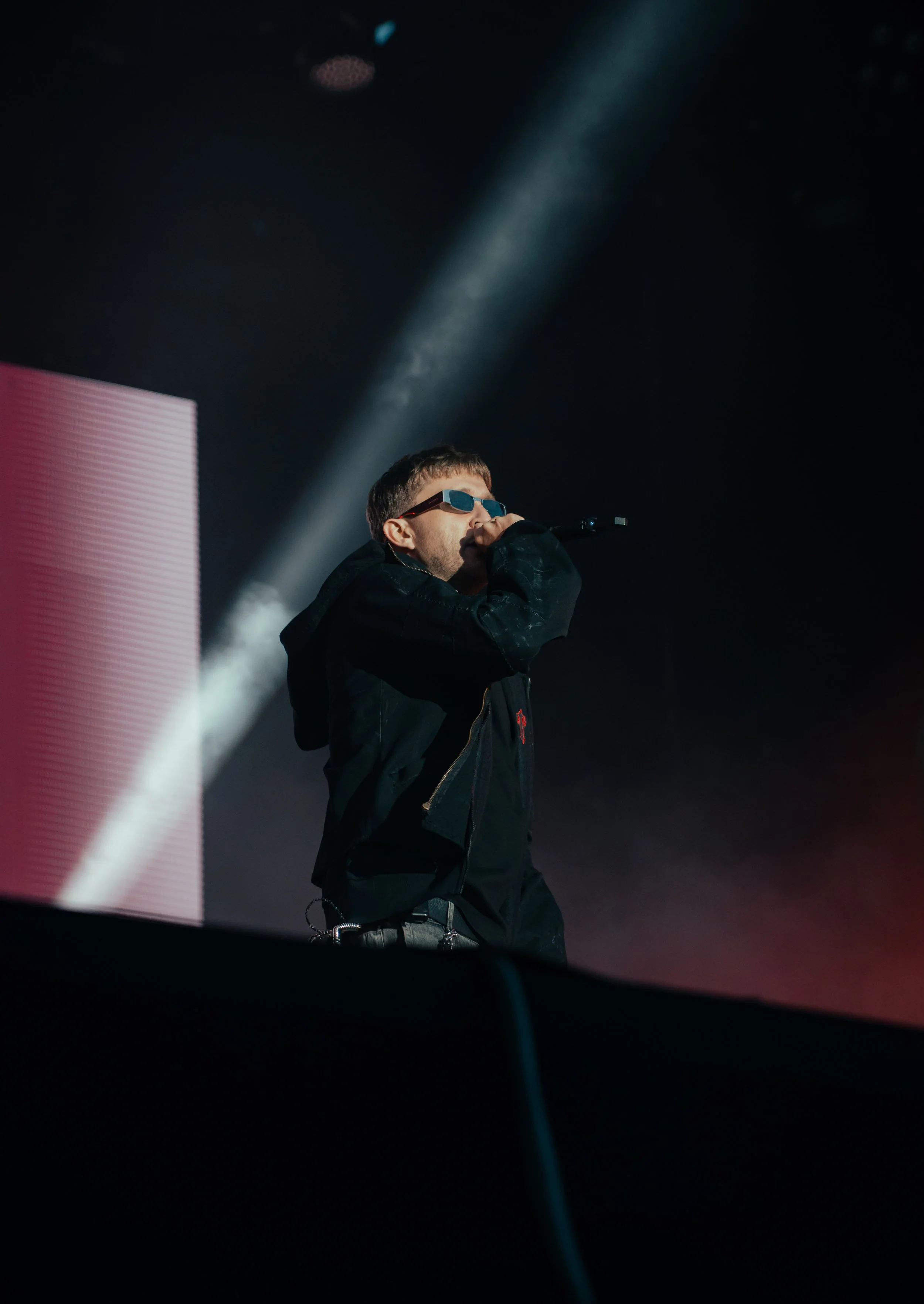 Un homme portant des lunettes de soleil noires se tient sur scène, utilisant un microphone, avec un éclairage de scène en arrière-plan.