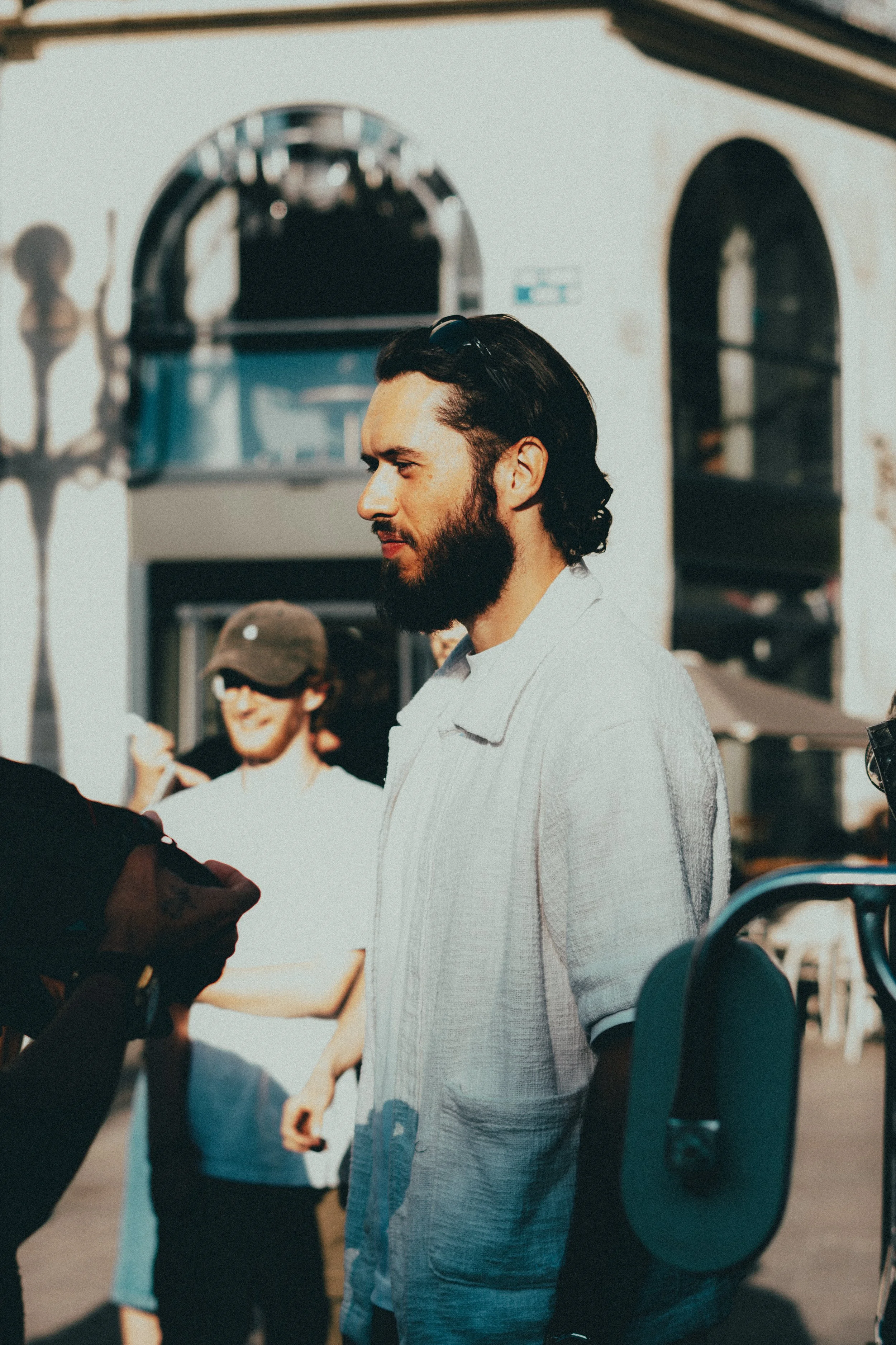 Un homme avec une barbe et des cheveux longs, portant une chemise claire, discutant avec quelqu'un dans une rue ensoleillée
