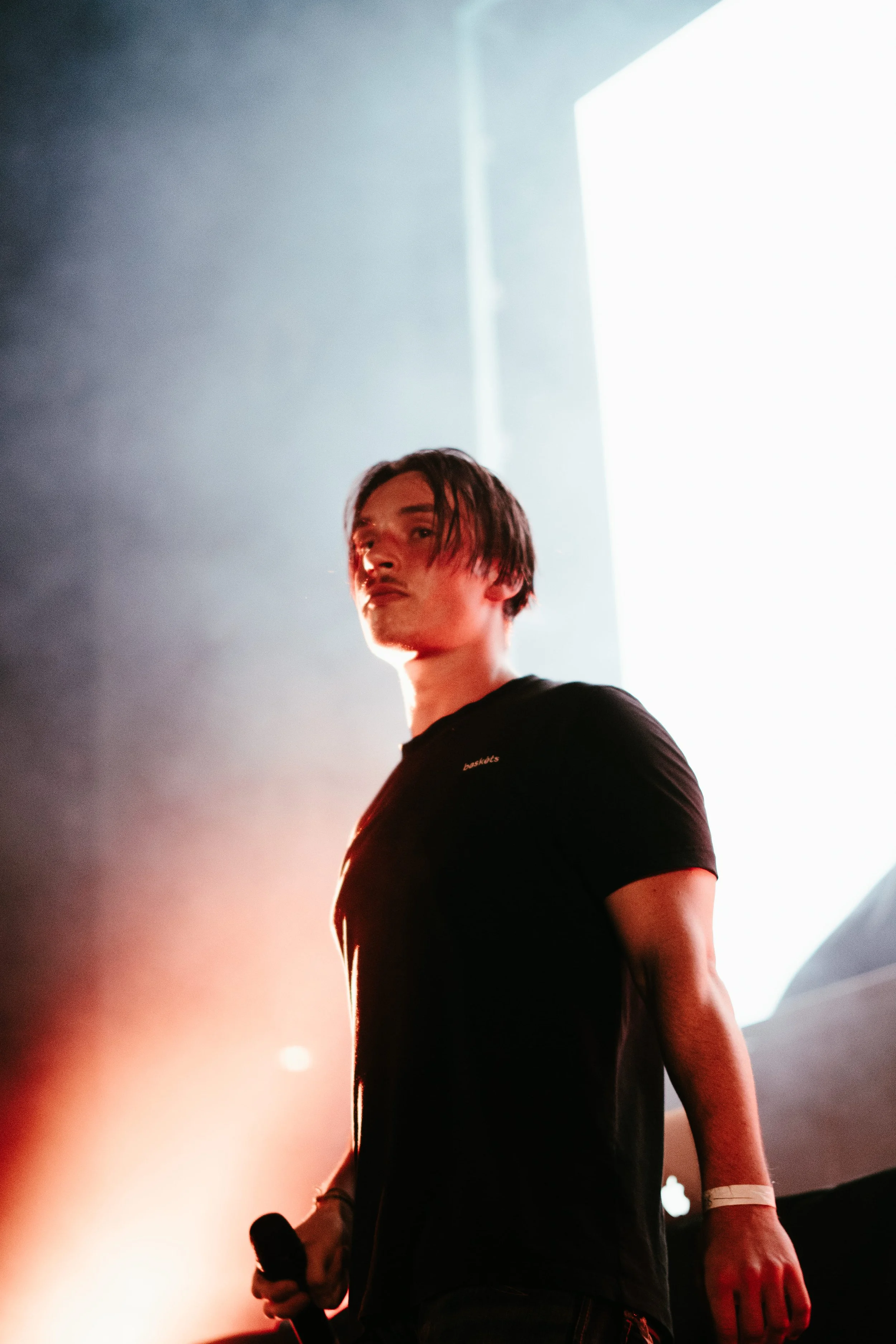 Un homme avec des cheveux courts et une barbe, tenant un microphone, se tient devant un fond lumineux.