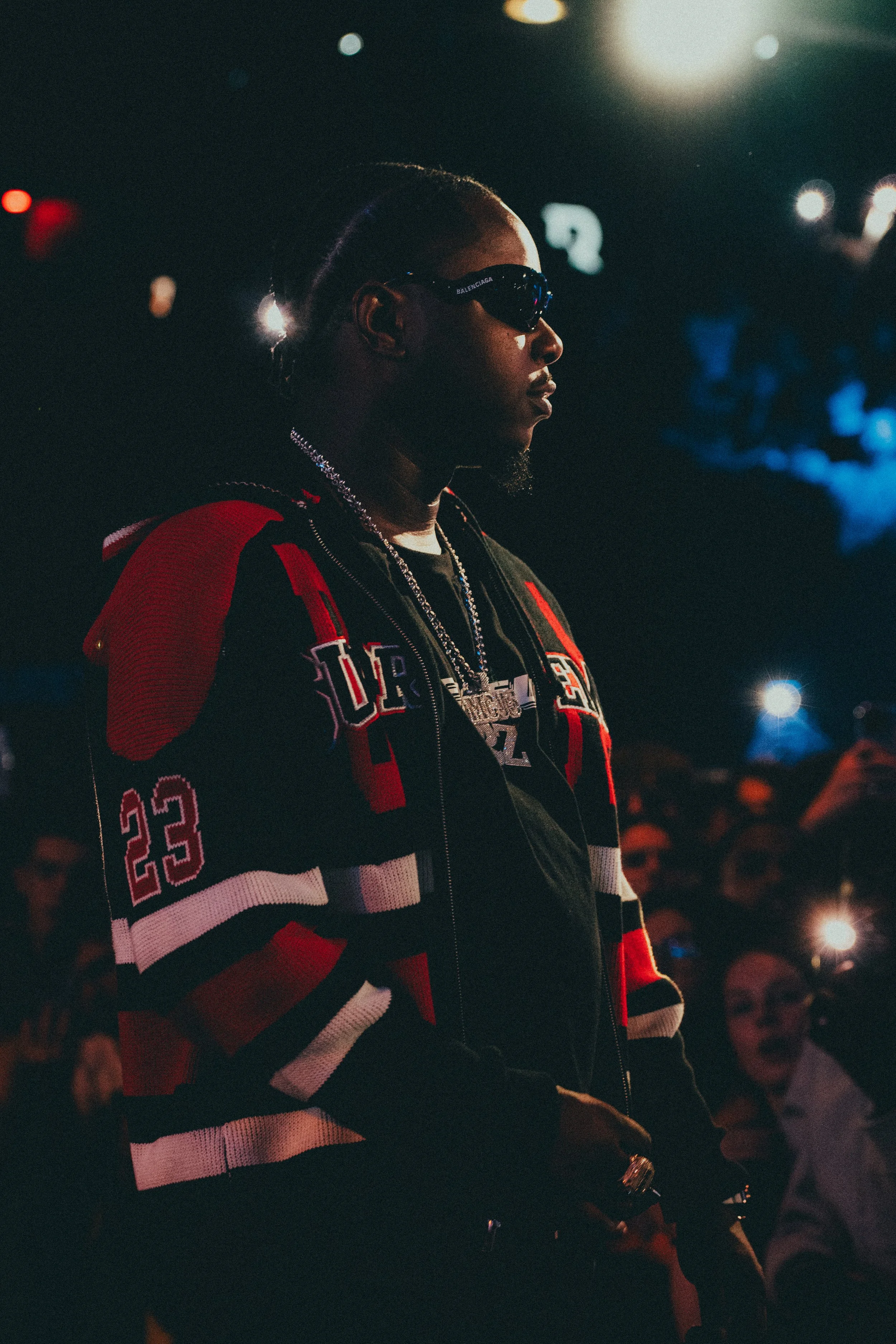 Un homme portant des lunettes de soleil, une veste de sport à rayures rouges, noires et blanches, et des chaînes en or, dans un lieu sombre avec un public en arrière-plan et des lumières brillantes.