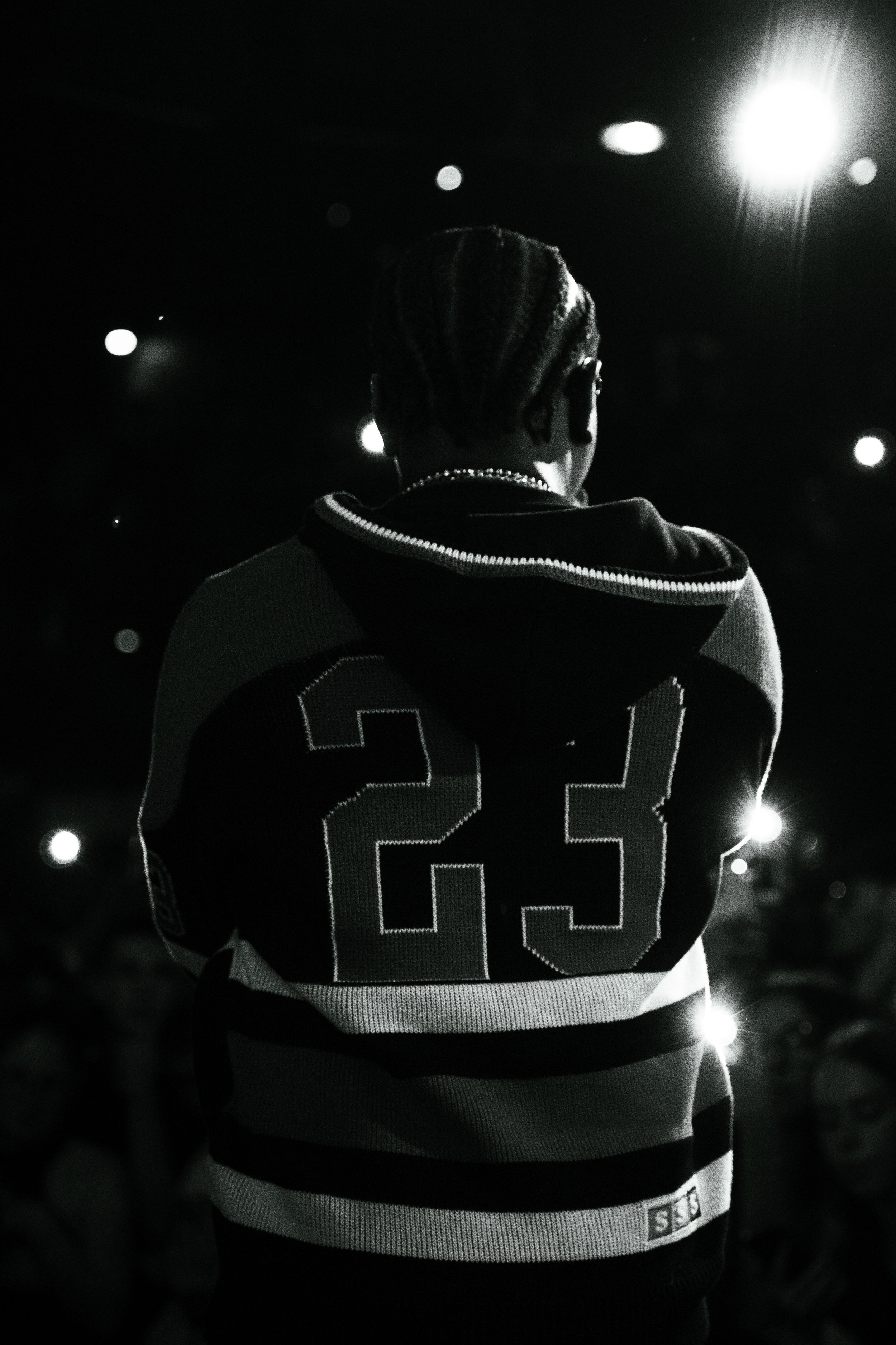 Personne vue de dos portant un sweat à capuche avec le numéro 23, dans un environnement nocturne avec des lumières brillantes et des reflets lumineux.