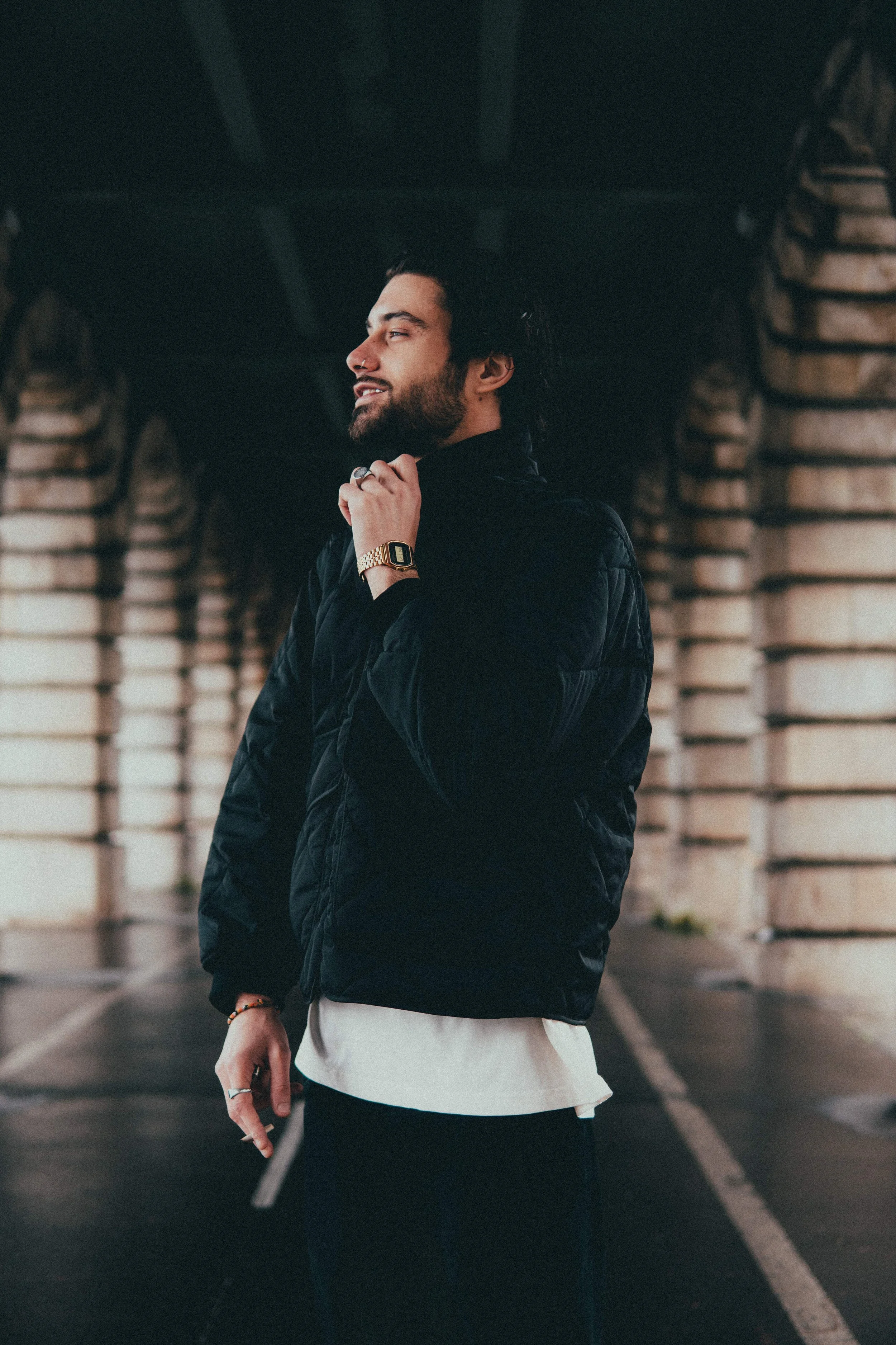 Un homme avec une barbe et un piercing, portant une montre dorée, se tient sous un pont en pierre, habillé d'une veste noire et d'un t-shirt blanc, dans un environnement urbain.