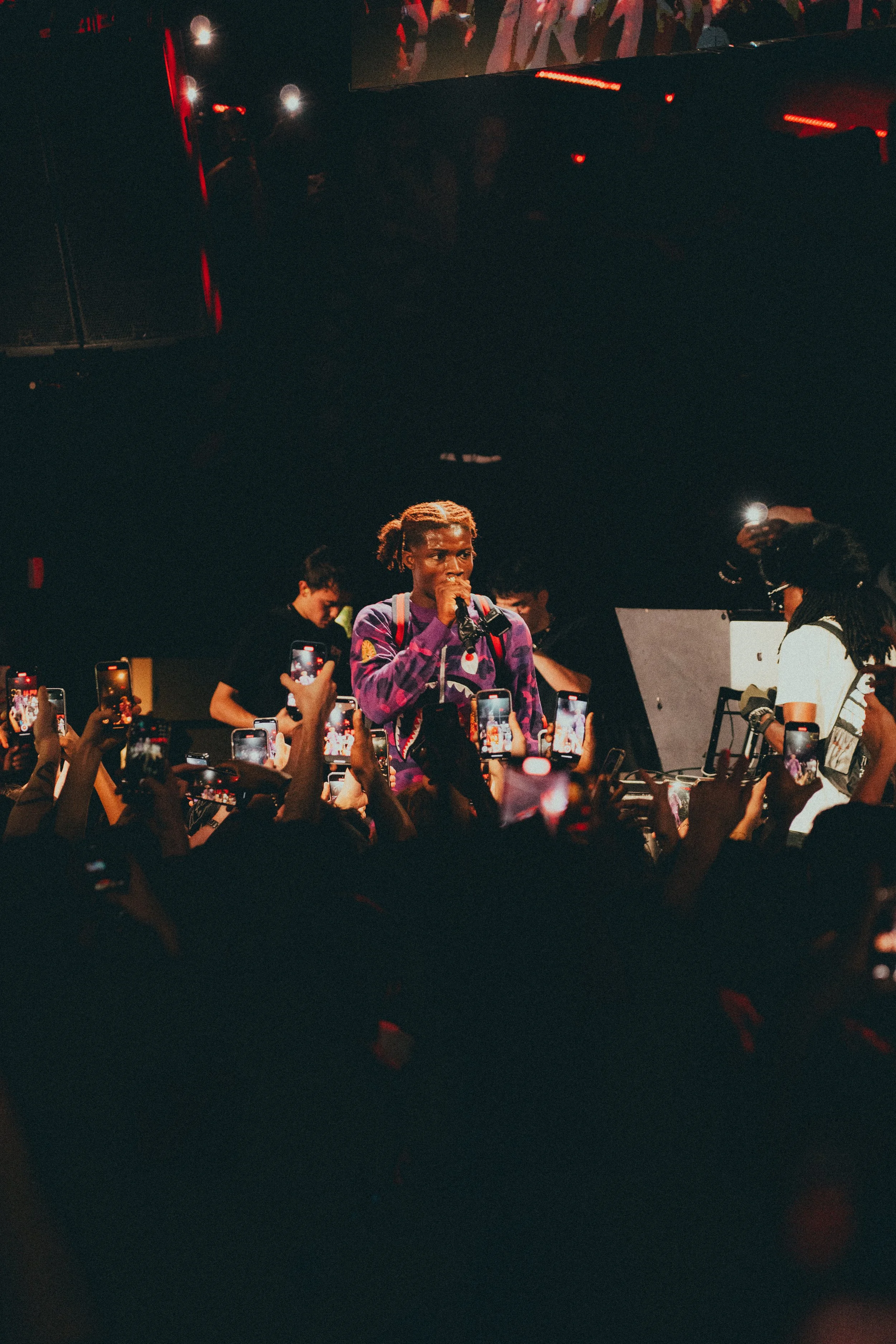 Un artiste ou DJ sur scène avec un microphone, entouré de nombreux spectateurs qui prennent des photos avec leurs téléphones.