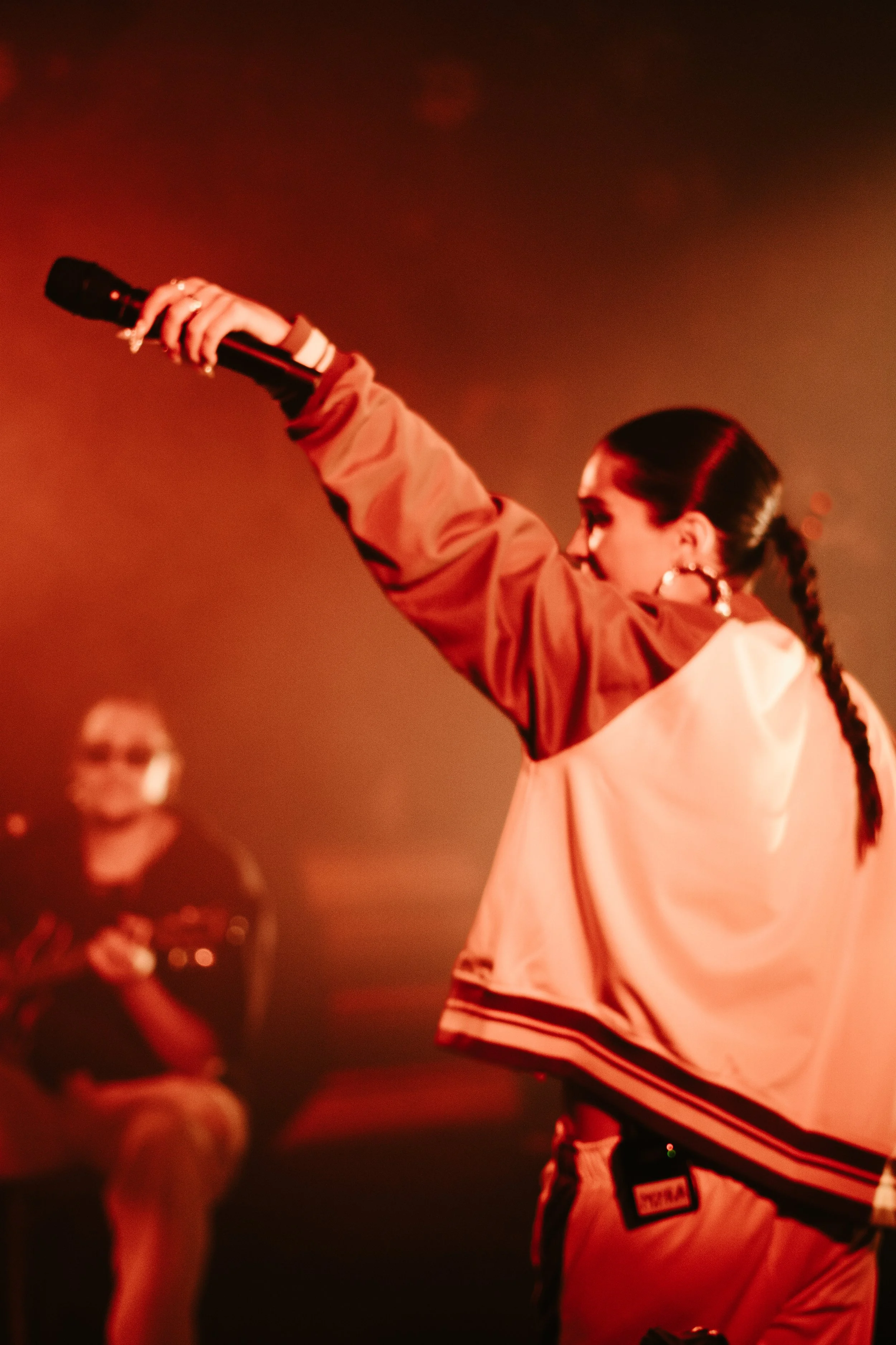 Femme sur scène, tenant un microphone, bras levé, avec une personne en fond qui joue de la guitare."