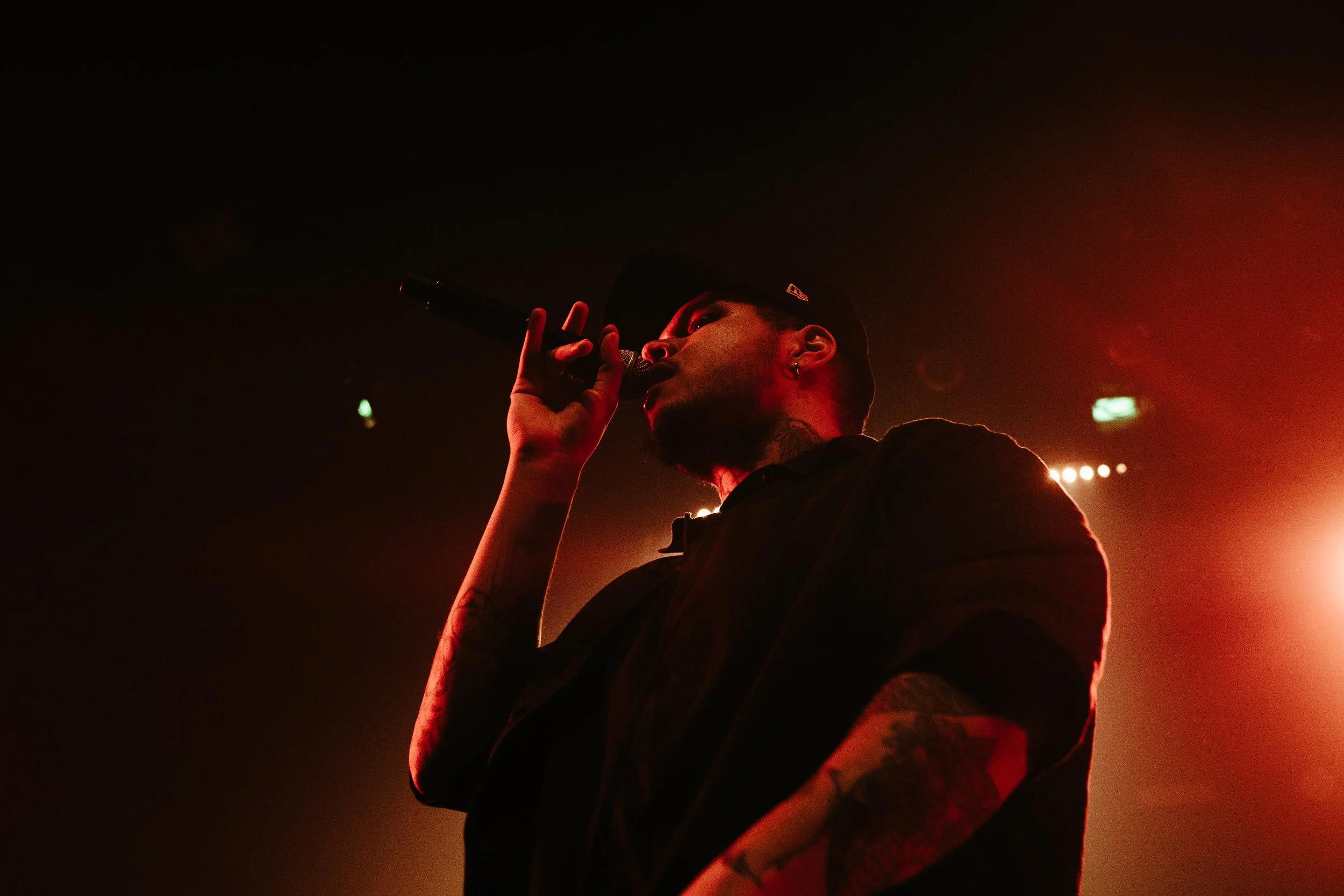 Jeune homme chantant dans un micro lors d'un concert, éclairage rouge et sombre en arrière-plan.