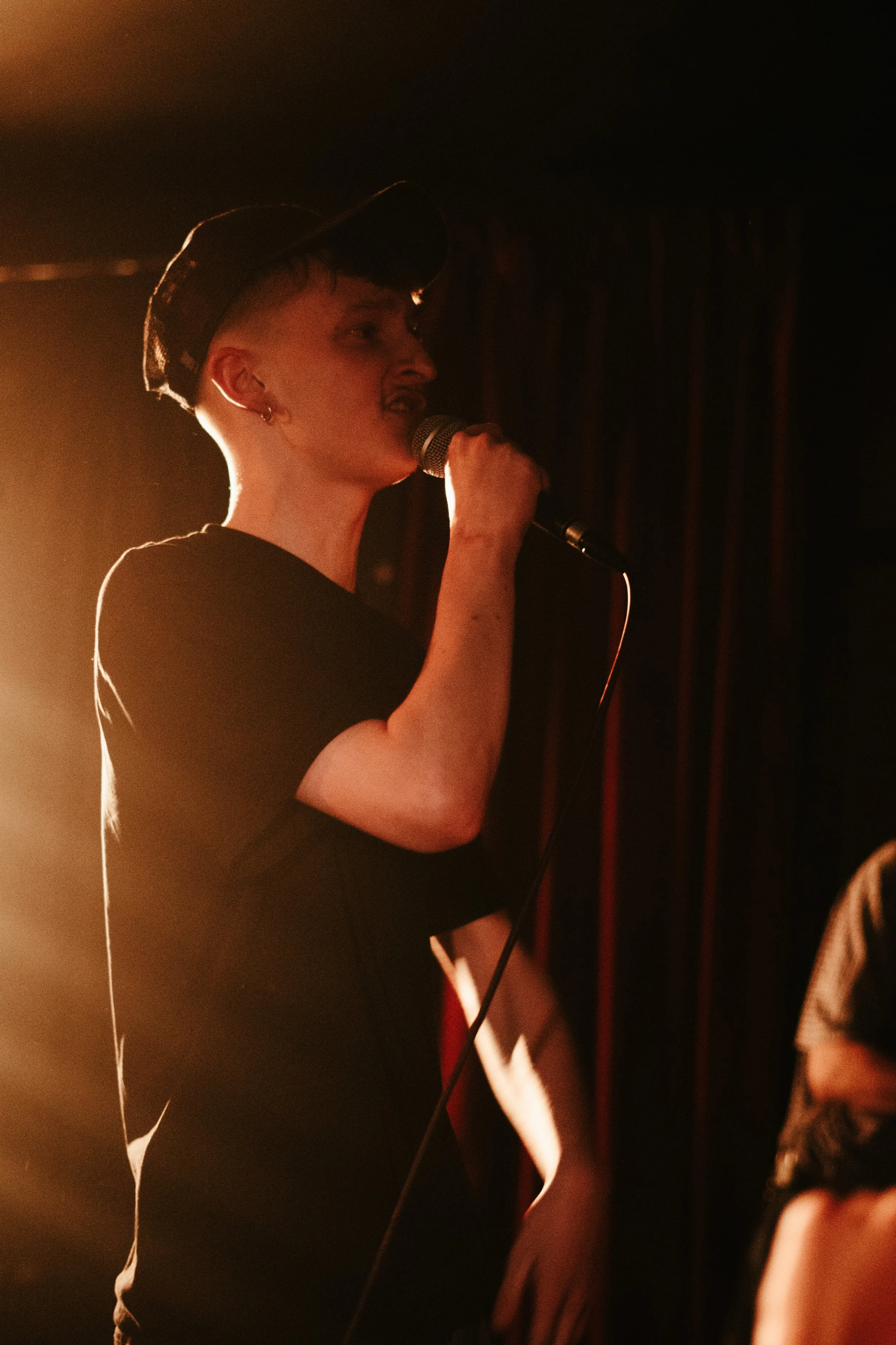 Une jeune femme avec une coupe de cheveux courte, portant une casquette, chante dans un micro, éclairée par une lumière chaude, en arrière-plan un rideau noir.