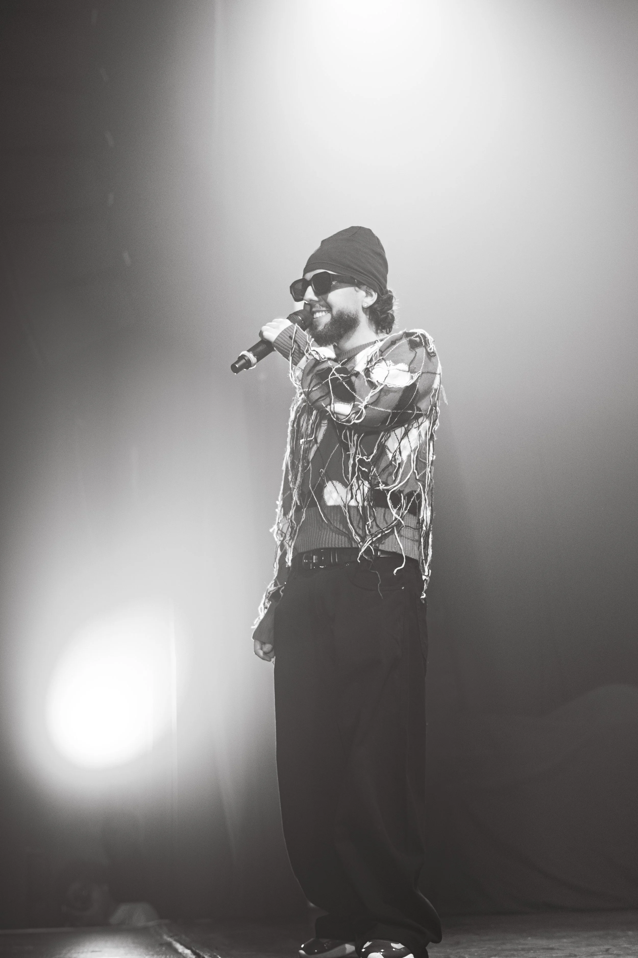 Un homme avec des lunettes de soleil et un bonnet, tenant un microphone en train de chanter ou parler sur scène, en noir et blanc.