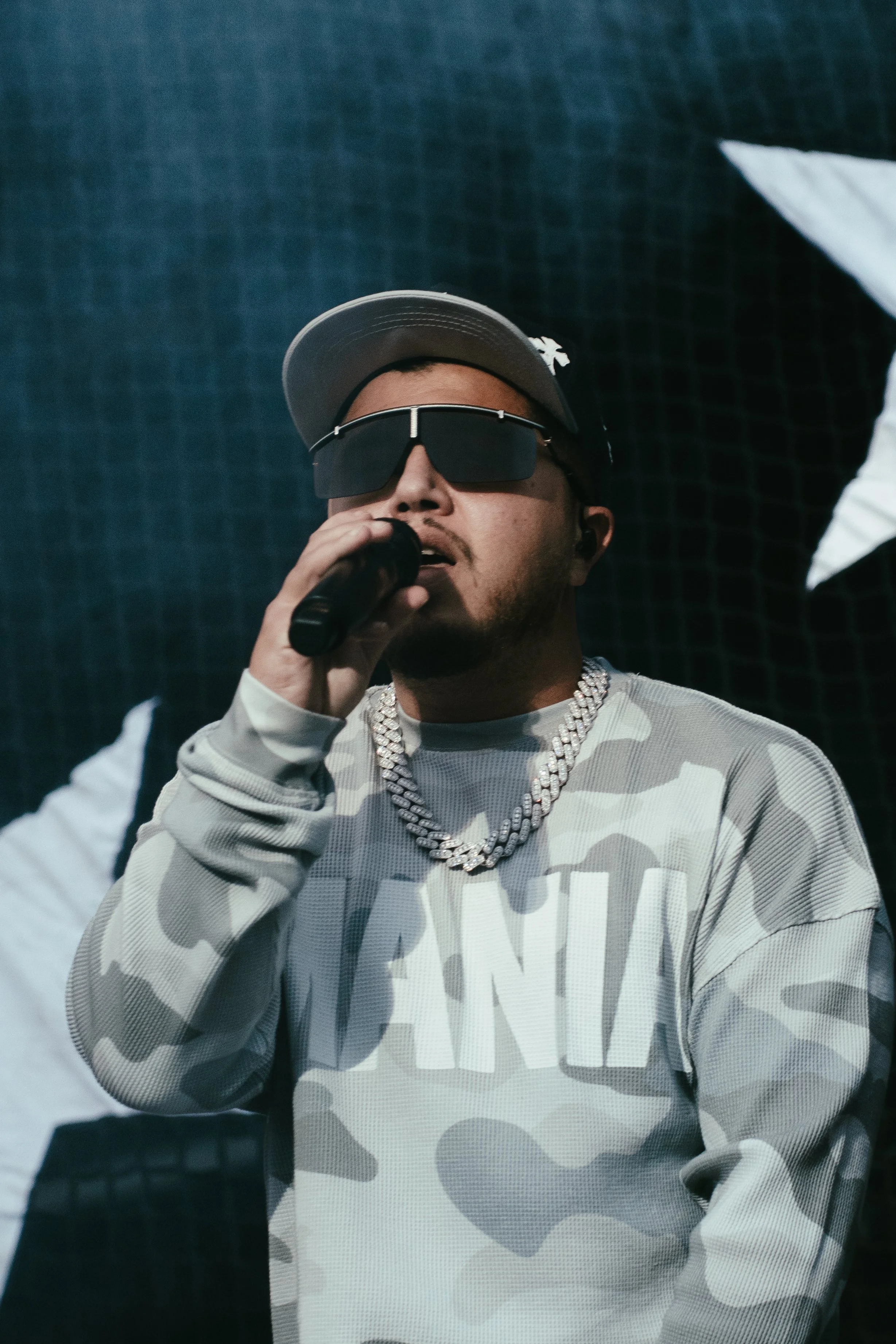Un rappeur portant des lunettes de soleil, une casquette, un maillot camouflage, un collier en or, tenant un microphone lors d'une performance musicale.