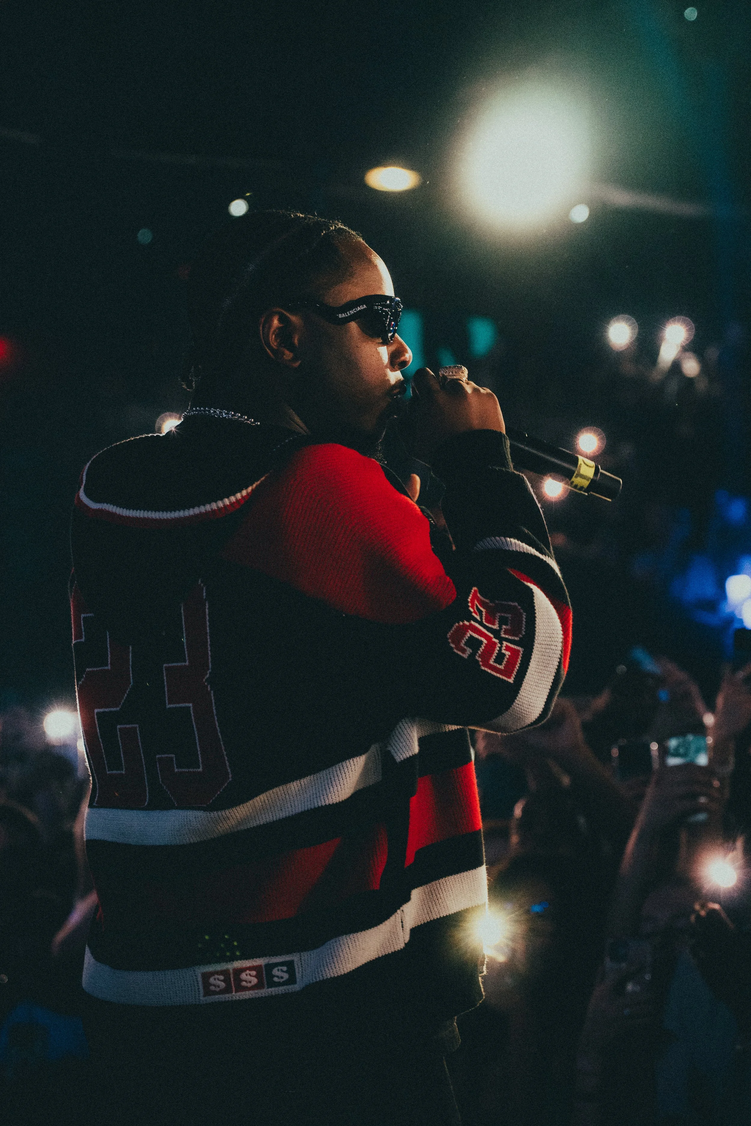 Un artiste portant des lunettes de soleil Nocturne en noir et rouge, tenant un micro, performe sur scène dans une ambiance sombre avec des lumières colorées.