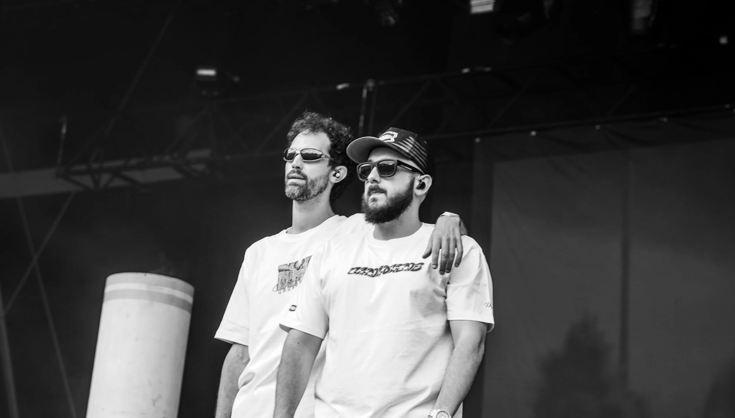Deux hommes portant des t-shirts blancs et des lunettes de soleil, un d'eux a un bras autour de l'autre, sur scène lors d'une performance musicale.