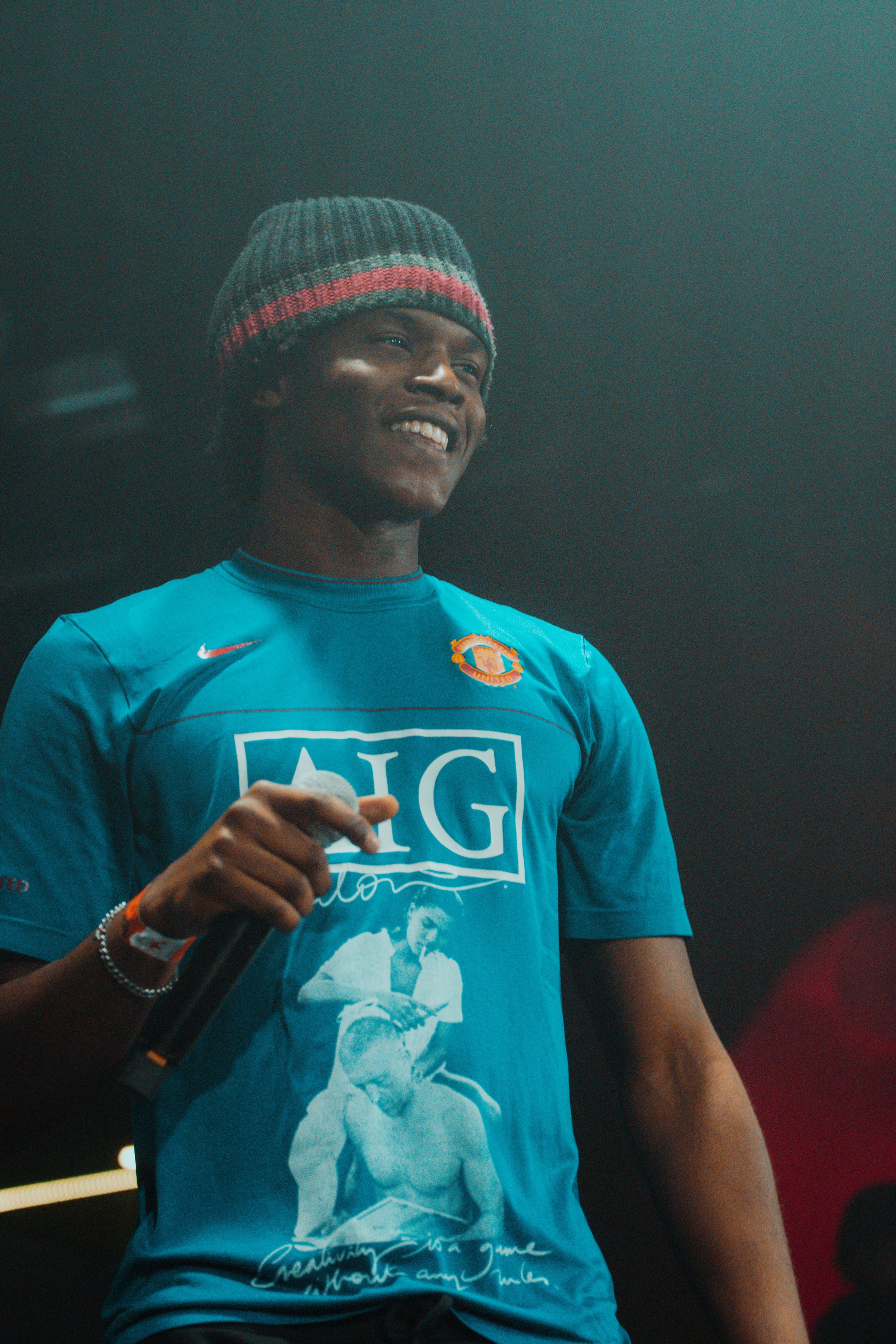 Jeune homme souriant portant un bonnet gris avec une bande rouge, un T-shirt bleu avec un logo Nike, une image et des inscriptions.