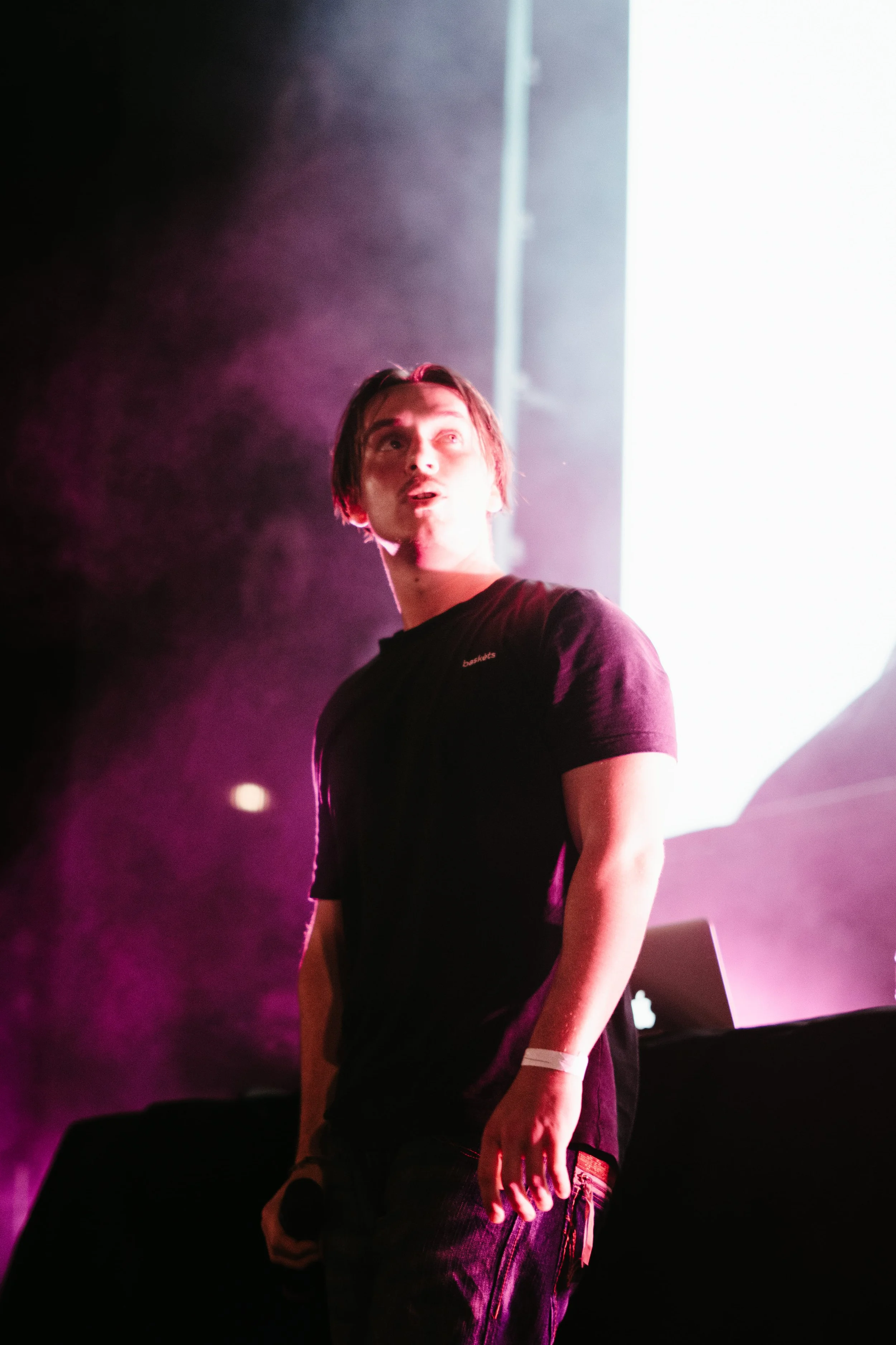 Un jeune homme en t-shirt noir sur scène, éclairé par une lumière blanche brillante en arrière-plan, avec des effets de fumée rose et violette.