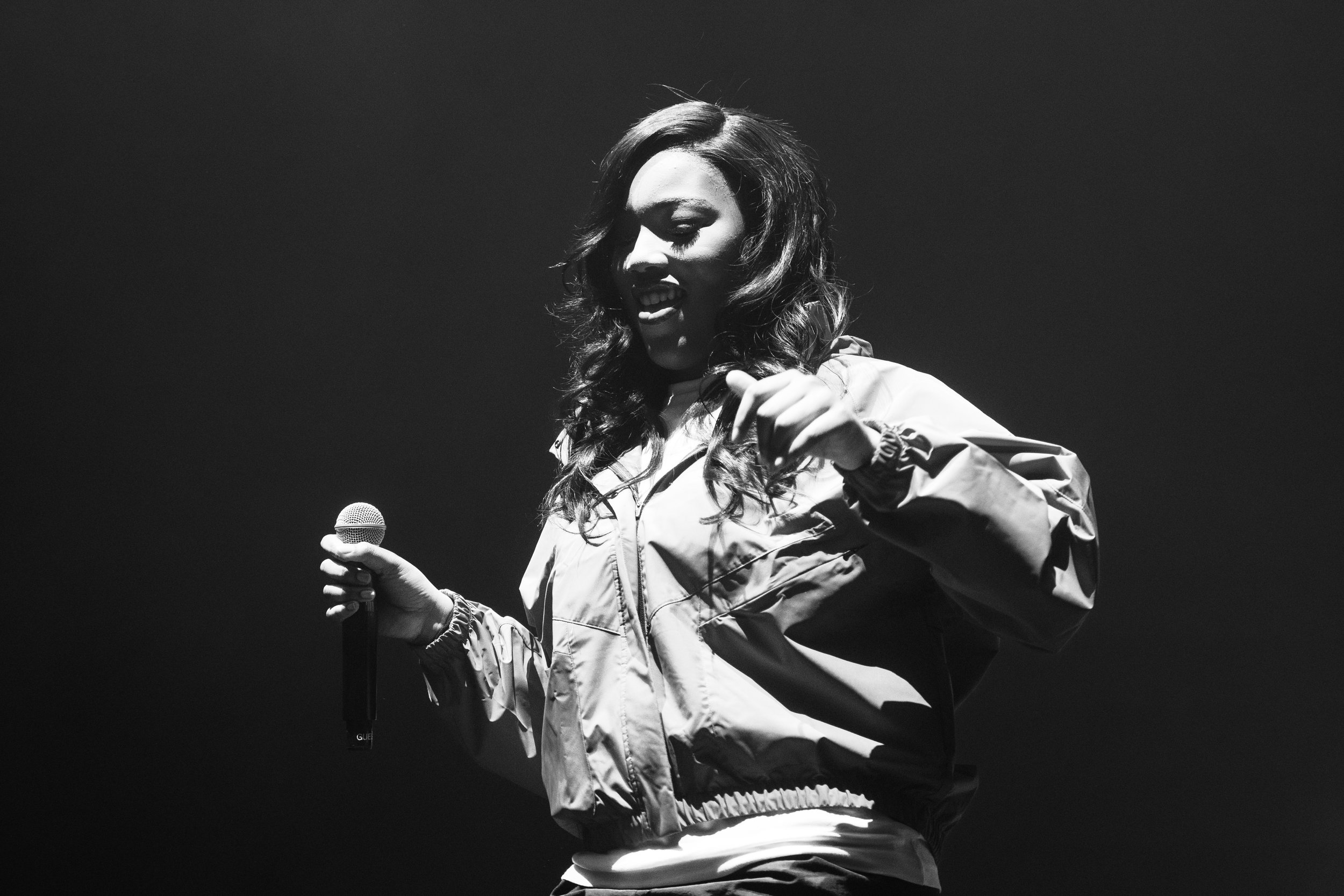 Femme tenant un microphone, parlant ou chantant sur scène, en noir et blanc.