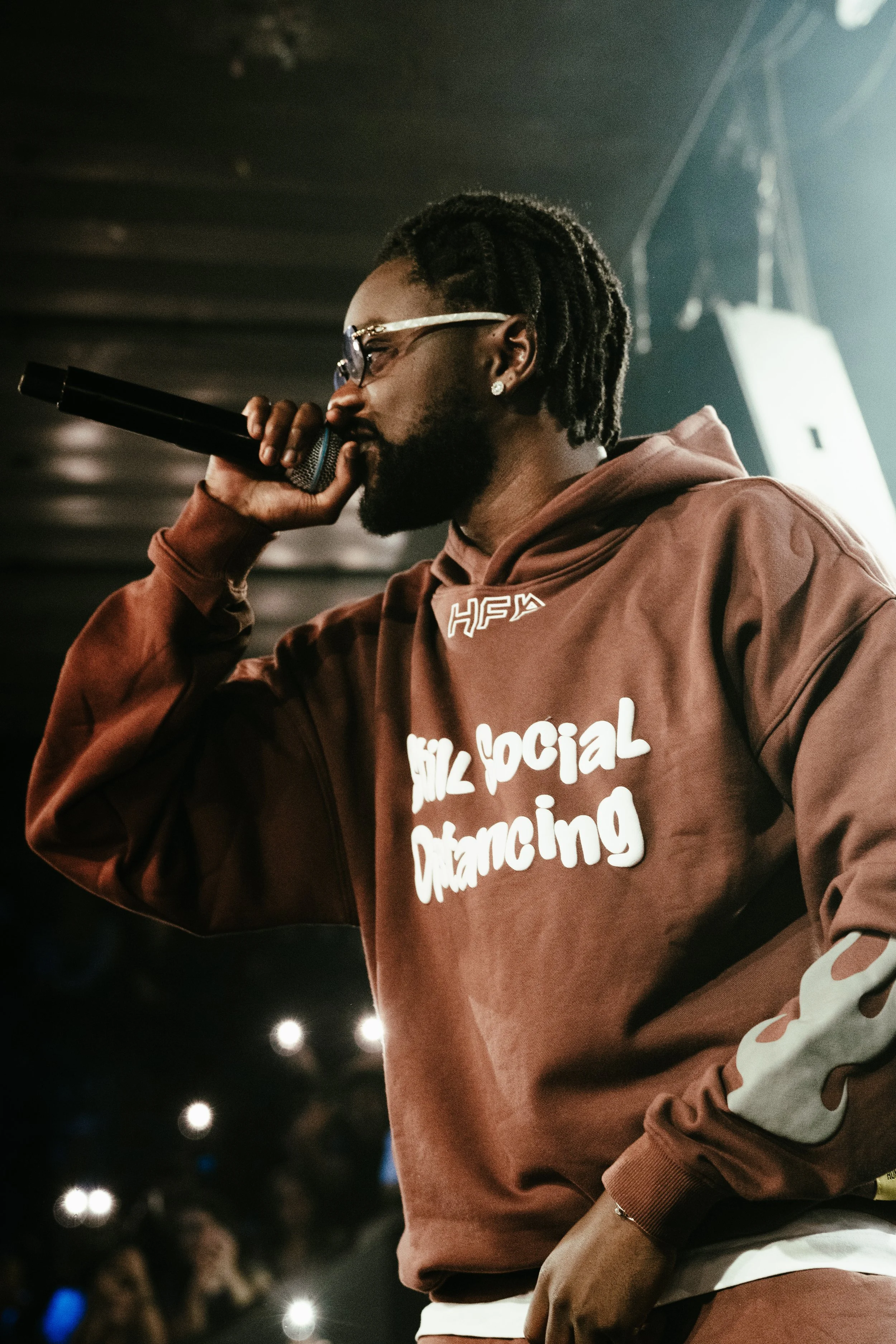 Un homme avec des dreadlocks, une barbe, portant des lunettes et un sweat à capuche marron avec l'inscription 'Social Dancing' en blanc, en train de chanter ou rapper lors d'une performance.