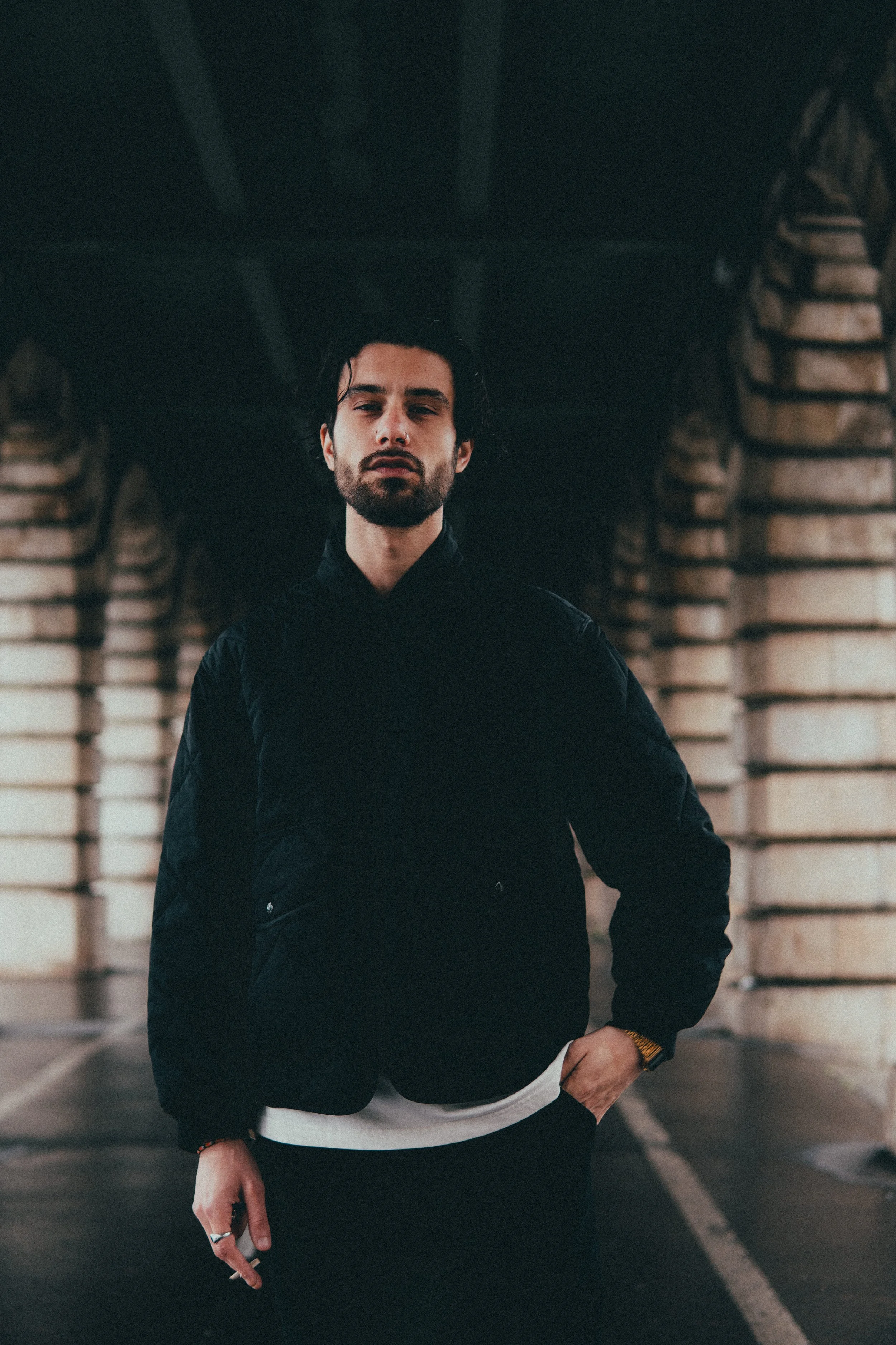 Un homme avec une barbe et des cheveux noirs court vêtue, portant une veste noire et une montre dorée, dans un parking souterrain avec des piliers en briques.