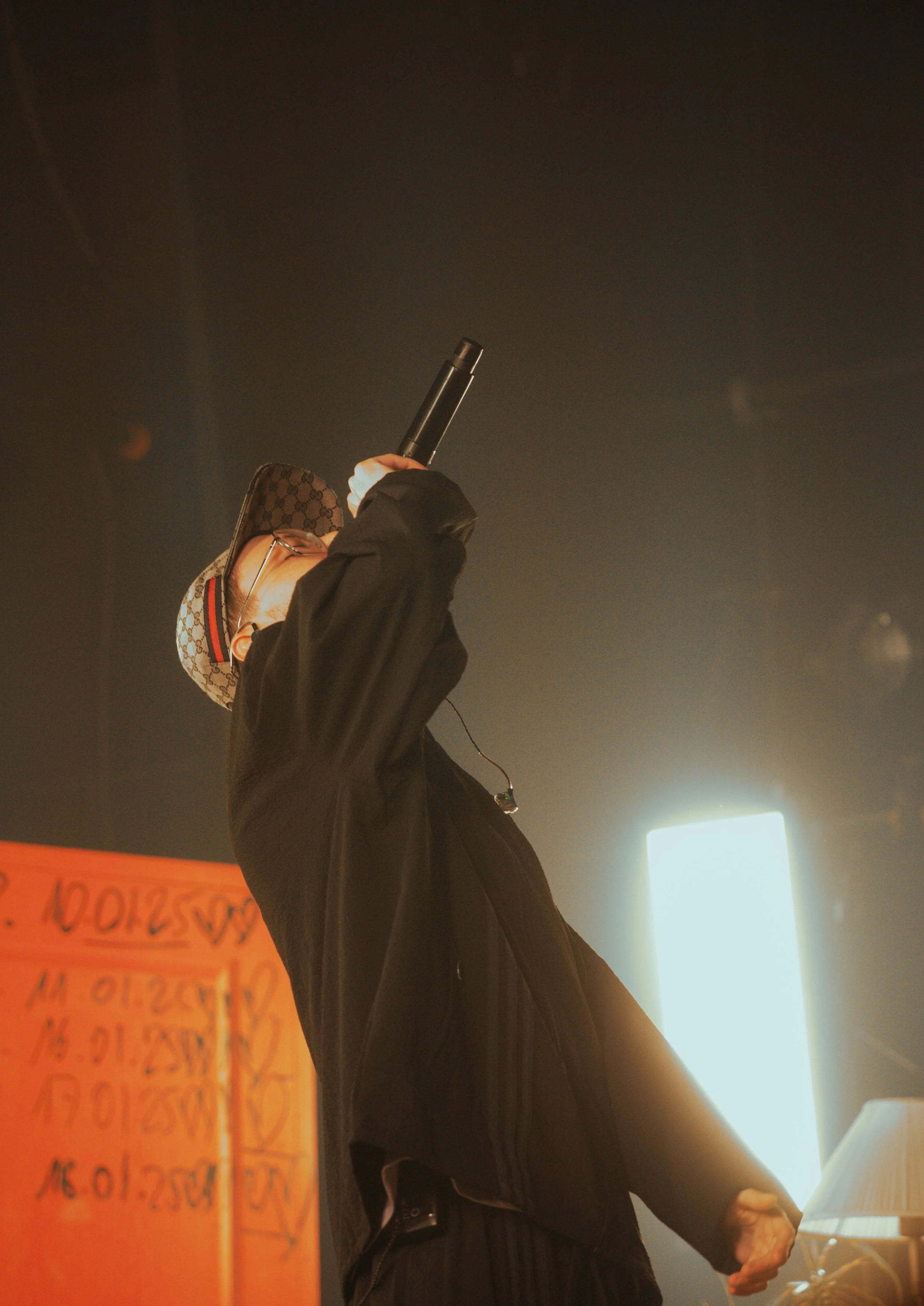 Homme portant un chapeau Gucci, portant des lunettes, chantant ou parlant dans un microphone, dans une scène sombre avec éclairage lumineux en arrière-plan.