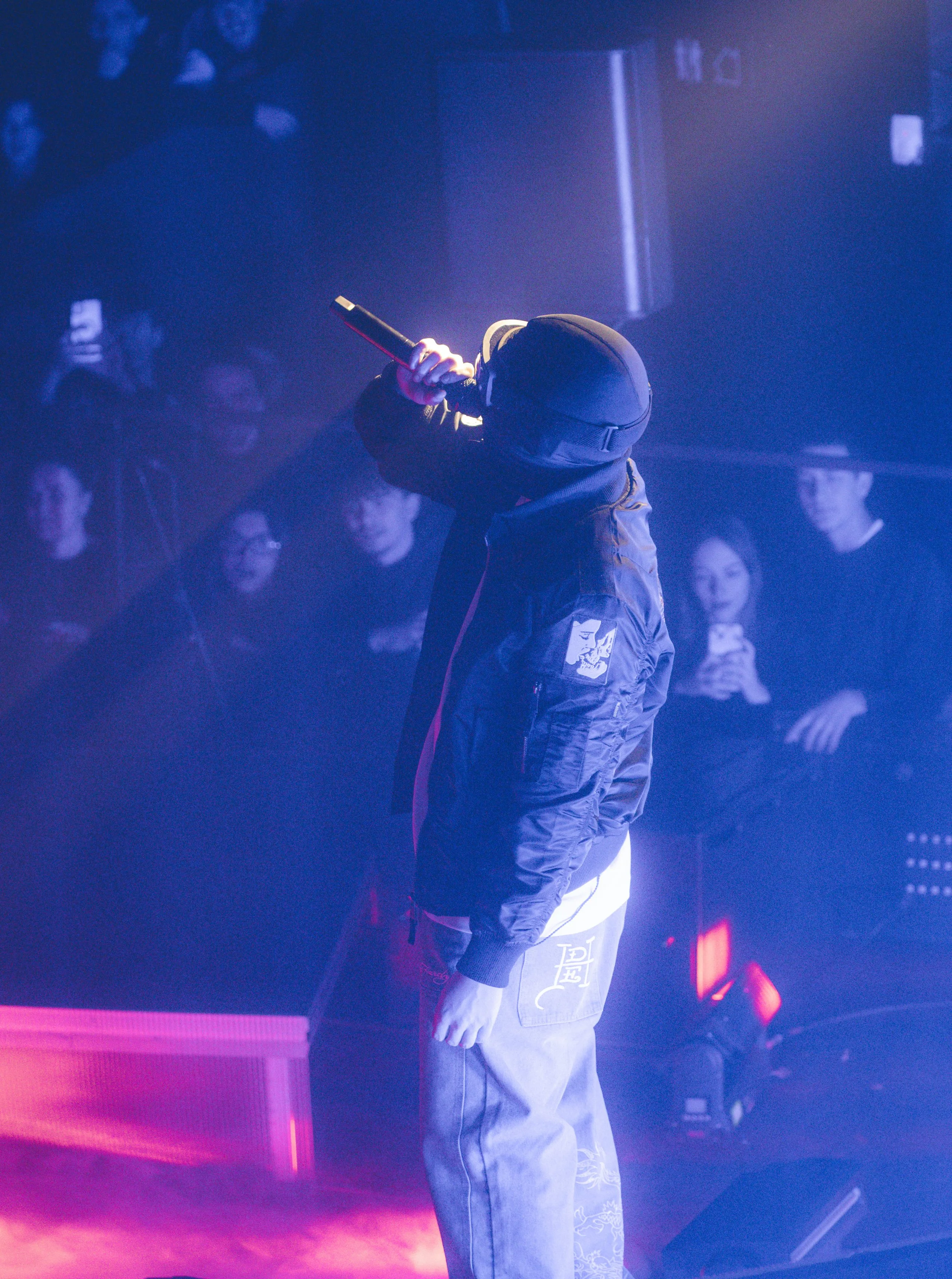 Un artiste en train de chanter ou de rapper sur scène, portant un casque et un blouson noir, avec un public en arrière-plan. Atmosphère sombre avec éclairage coloré.