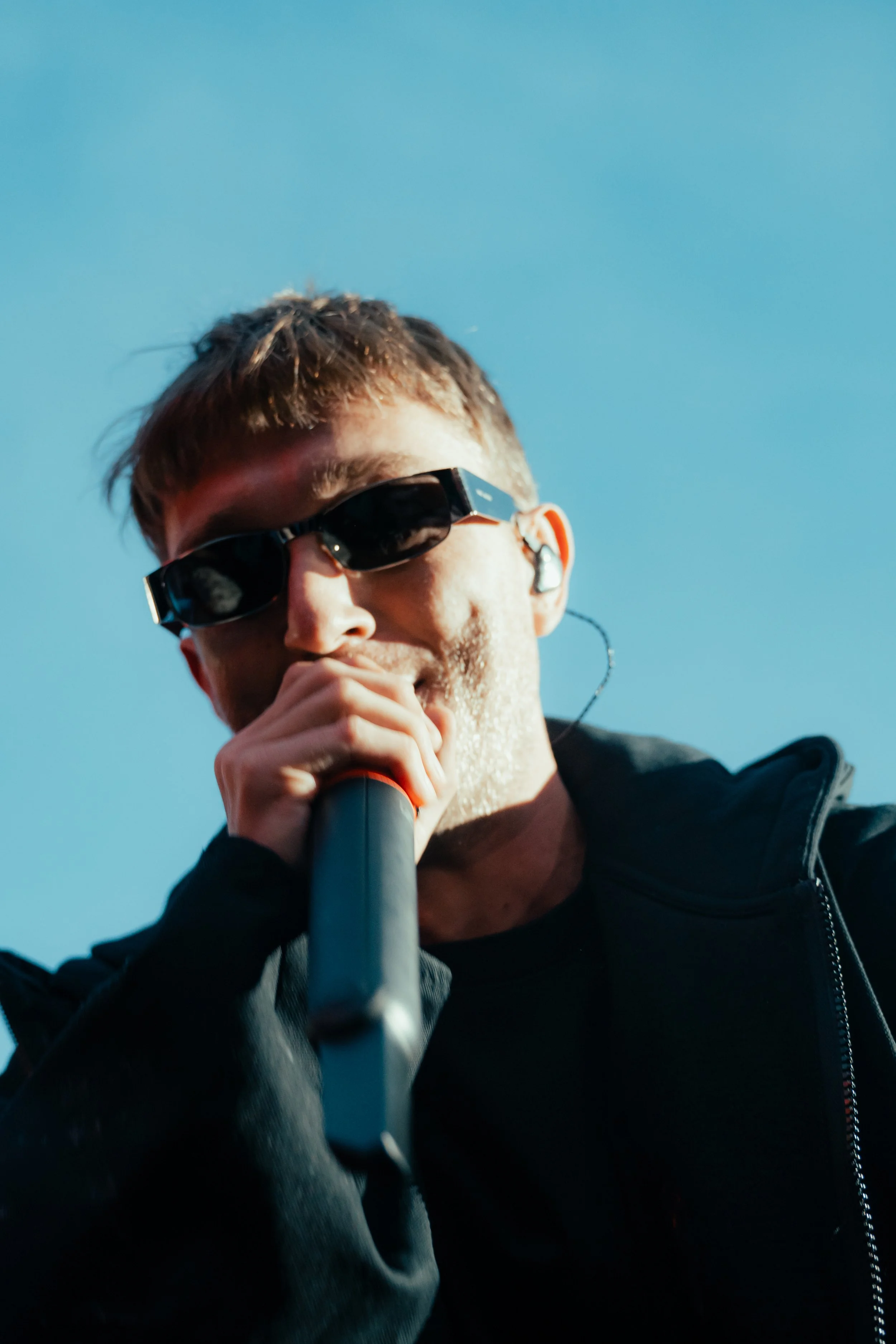 Un jeune homme portant des lunettes de soleil noires, tenant un microphone et portant un écouteur, en train de chanter ou de parler contre un ciel bleu clair.