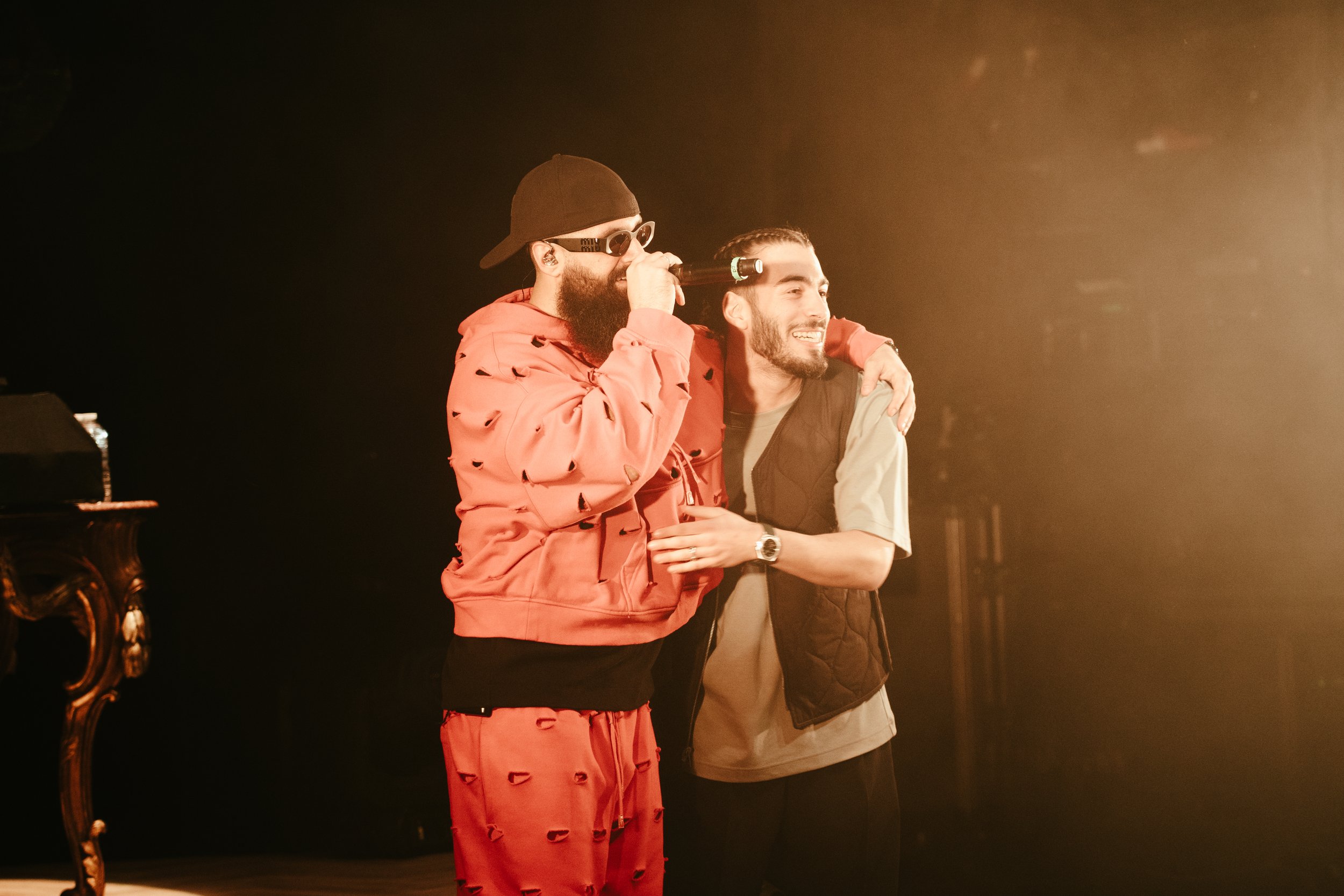 Deux hommes souriants, l'un avec une barbe et lunettes de soleil portant une veste rouge, et l'autre avec une barbe portant un t-shirt beige et un gilet noir, se tiennent ensemble dans une scène lumineuse, semblant partager un moment convivial.