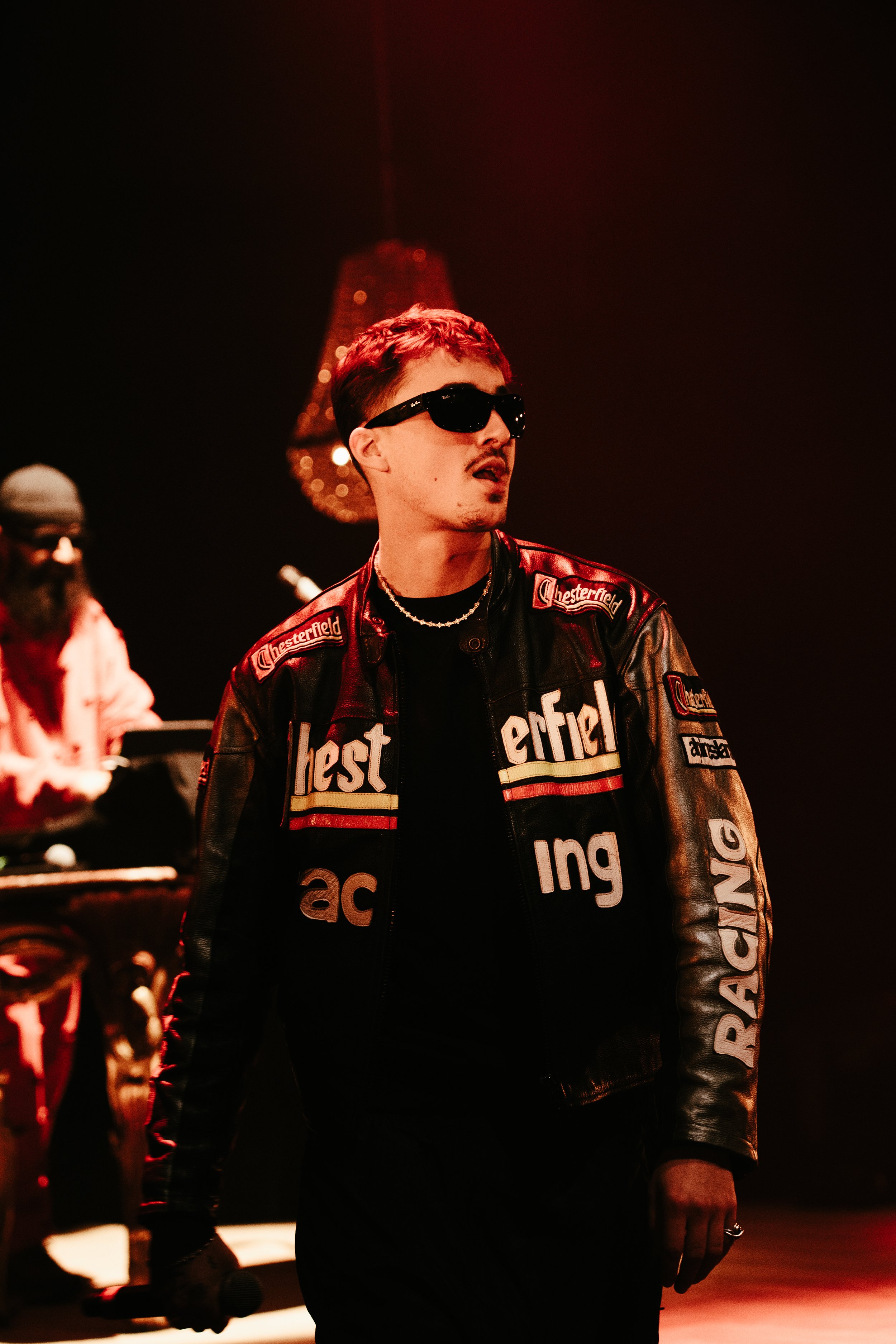 Un homme avec des lunettes de soleil noires portant une veste en cuir noir avec des logos 'Chesterfield' et 'Pack' sur scène, avec un éclairage rouge et une personne en arrière-plan utilisant un clavier.