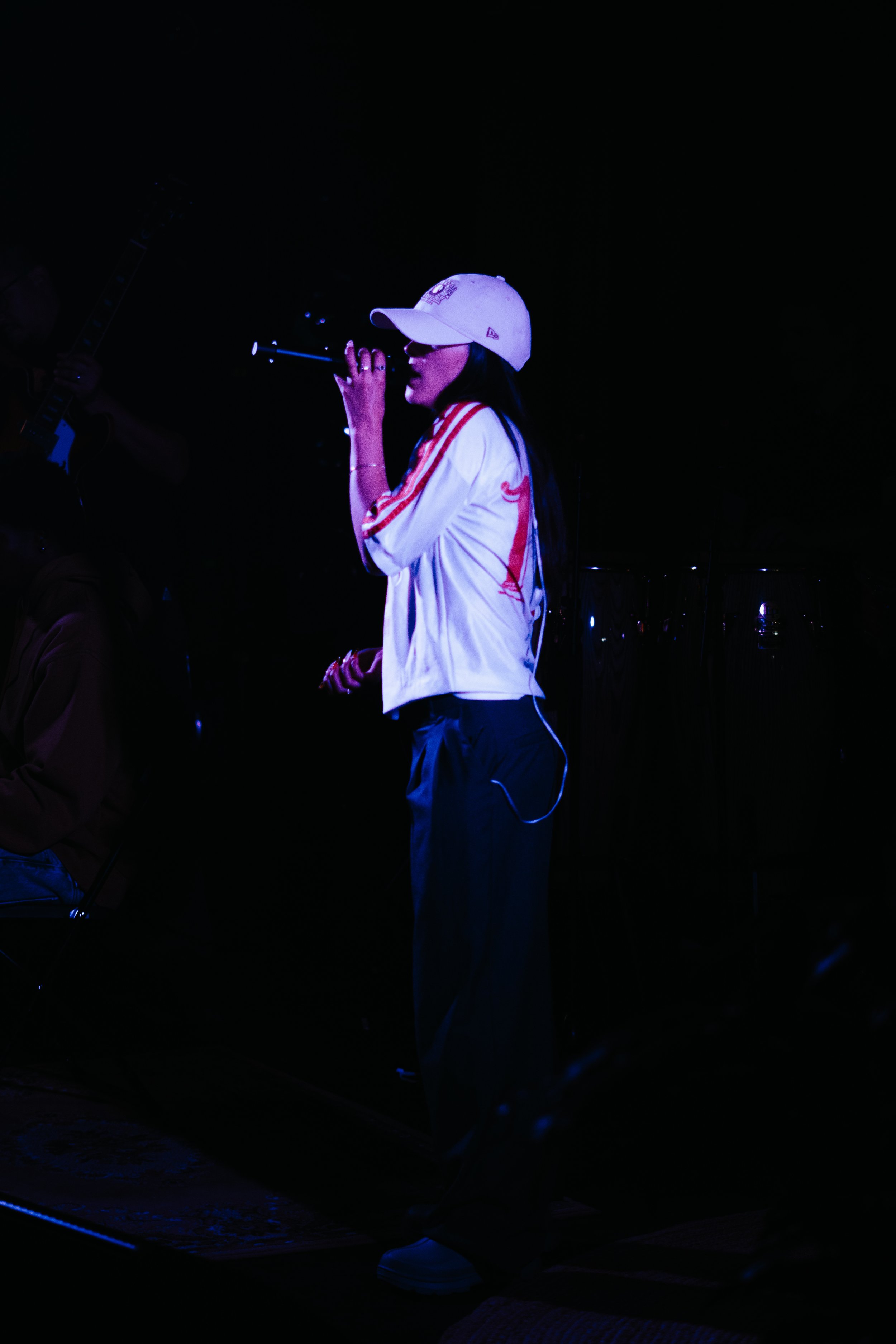 Une femme chante dans un microphone lors d'un spectacle, portant une casquette blanche et un survêtement blanc avec des rayures rouges, dans une ambiance sombre et colorée.