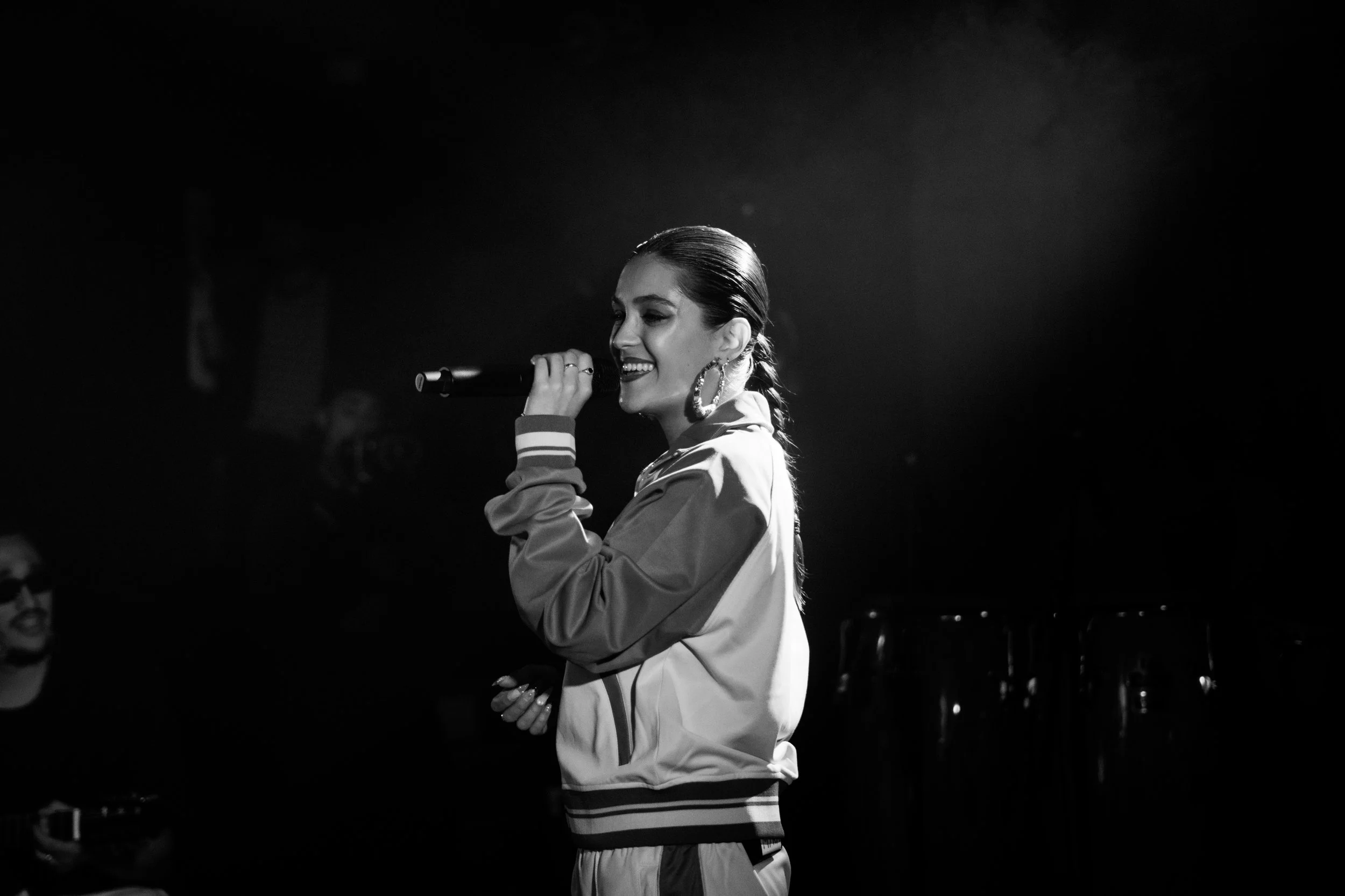 Une femme souriante avec des boucles d'oreilles grands, tenant un micro en scène dans un environnement sombre.