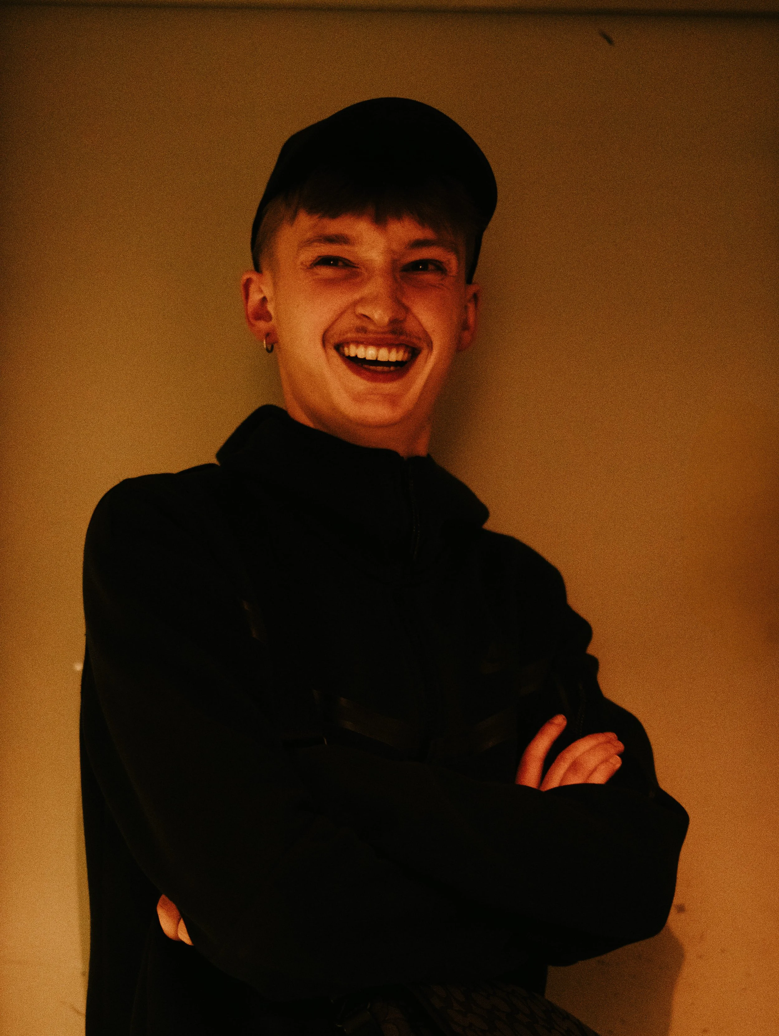 Un jeune homme souriant avec un casque et un sweat noir, croisant les bras, debout contre un mur beige.