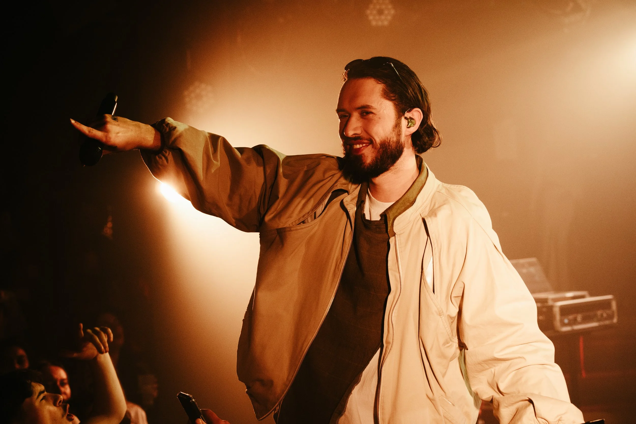 Homme souriant avec une barbe, portant un casque auditif, tenant un micro et faisant un signe aux spectateurs lors d'une performance sur scène.