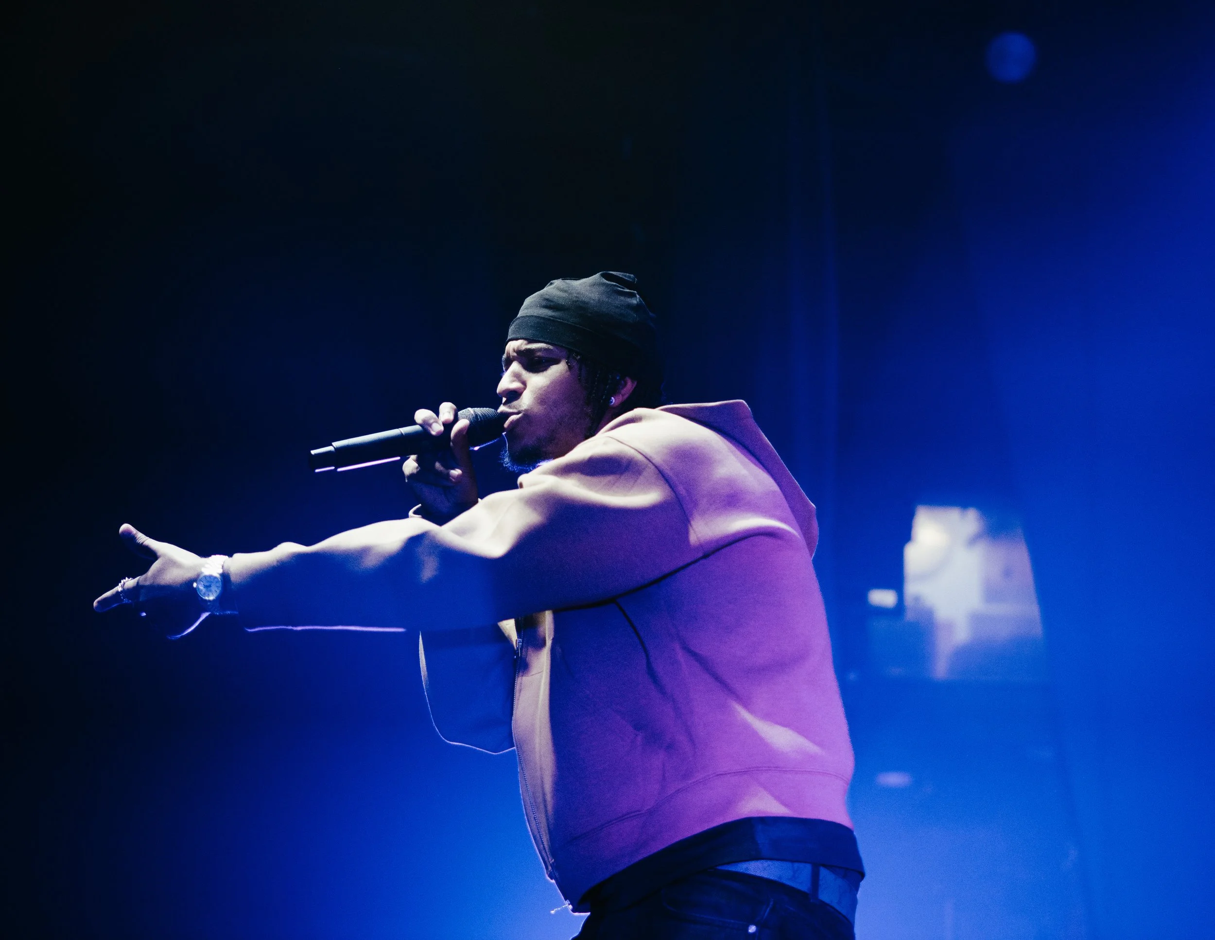 Un homme en sweat à capuche beige et bonnet noir, chantant dans un micro, sous un éclairage bleu en scène.