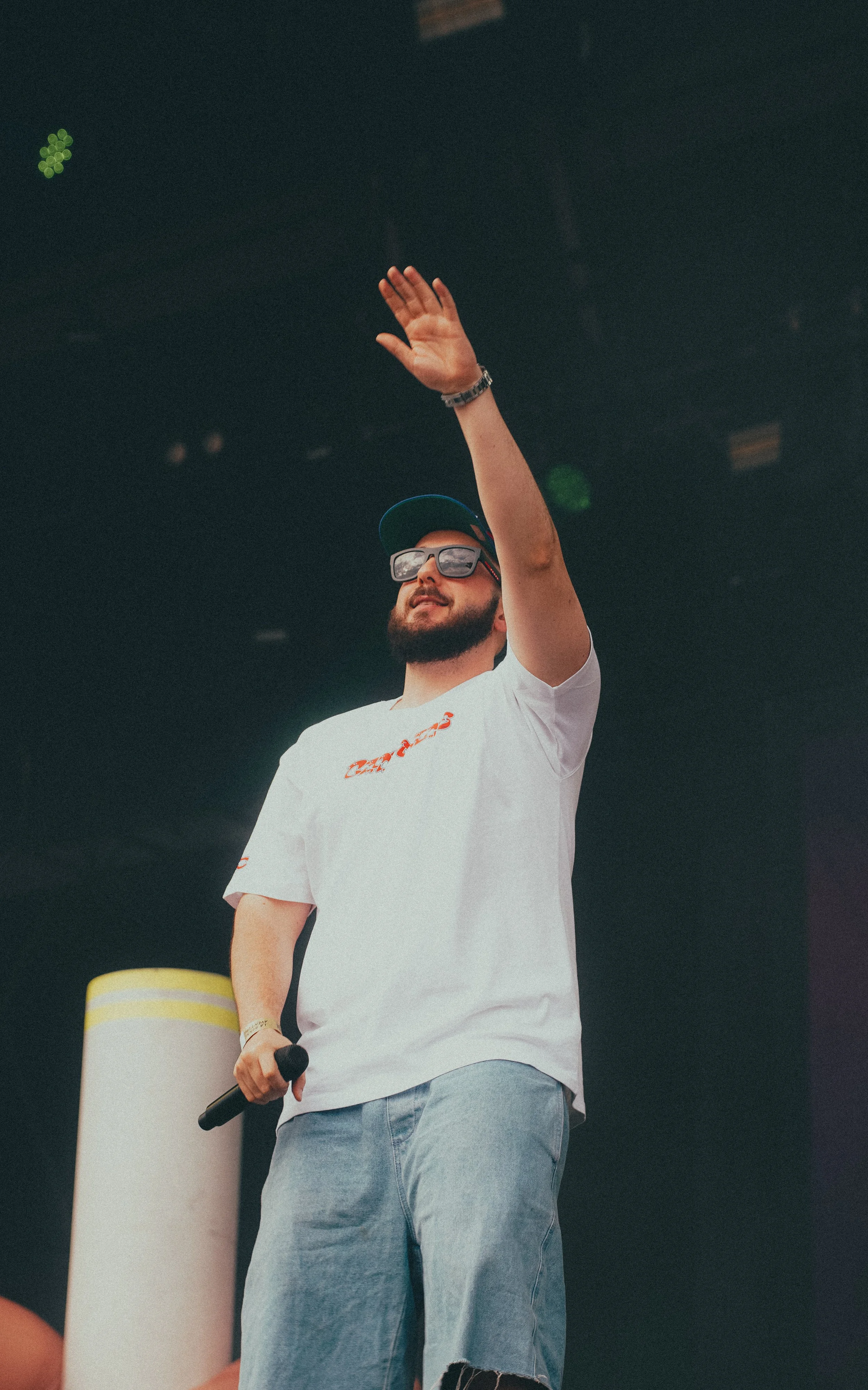 Un homme portant une casquette, des lunettes de soleil et un t-shirt blanc joue avec un micro lors d'un évènement en intérieur.