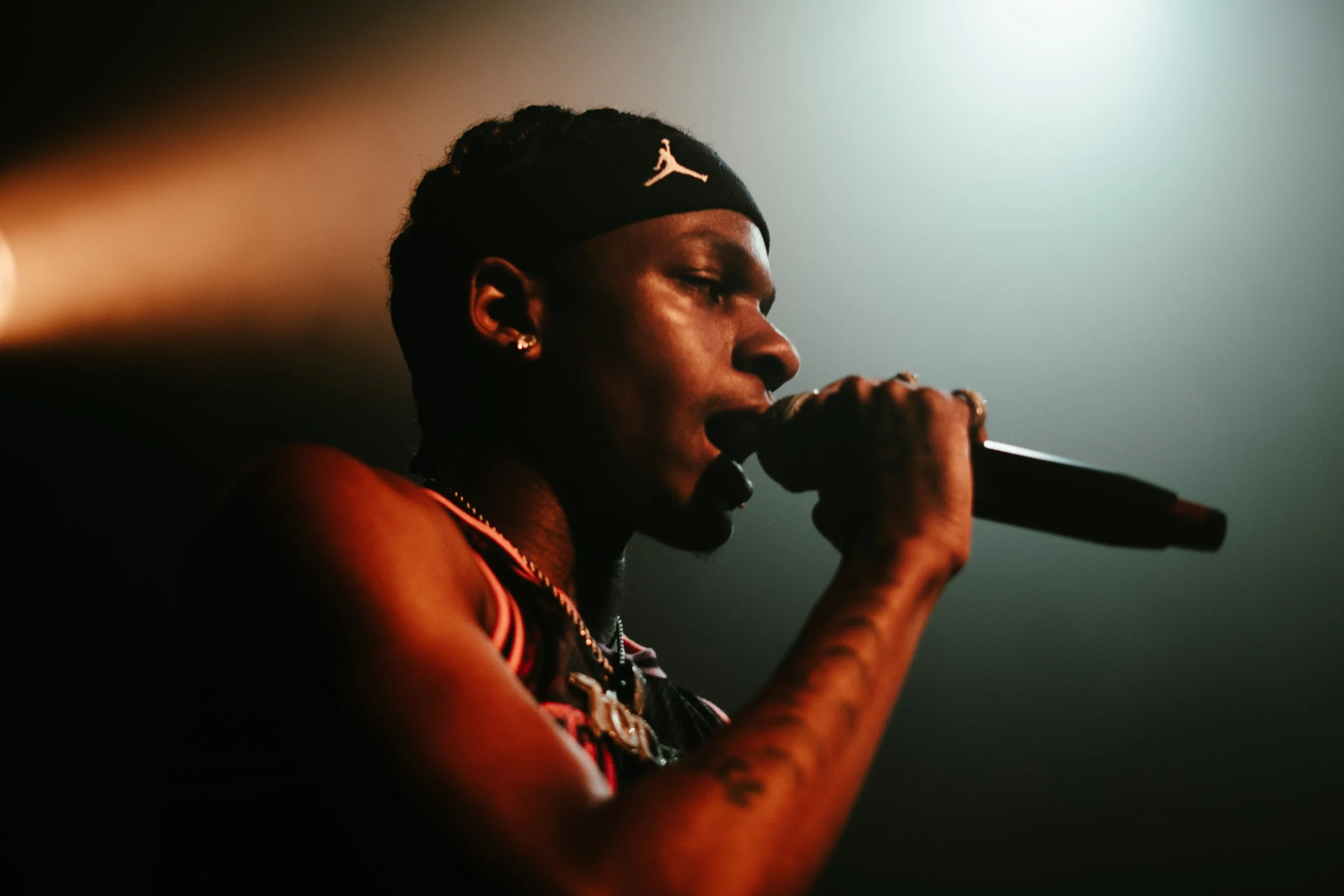 Jeune homme noir chantant dans un micro, portant un bandeau noir avec un logo blanc, en mode sombre avec un fond flou.