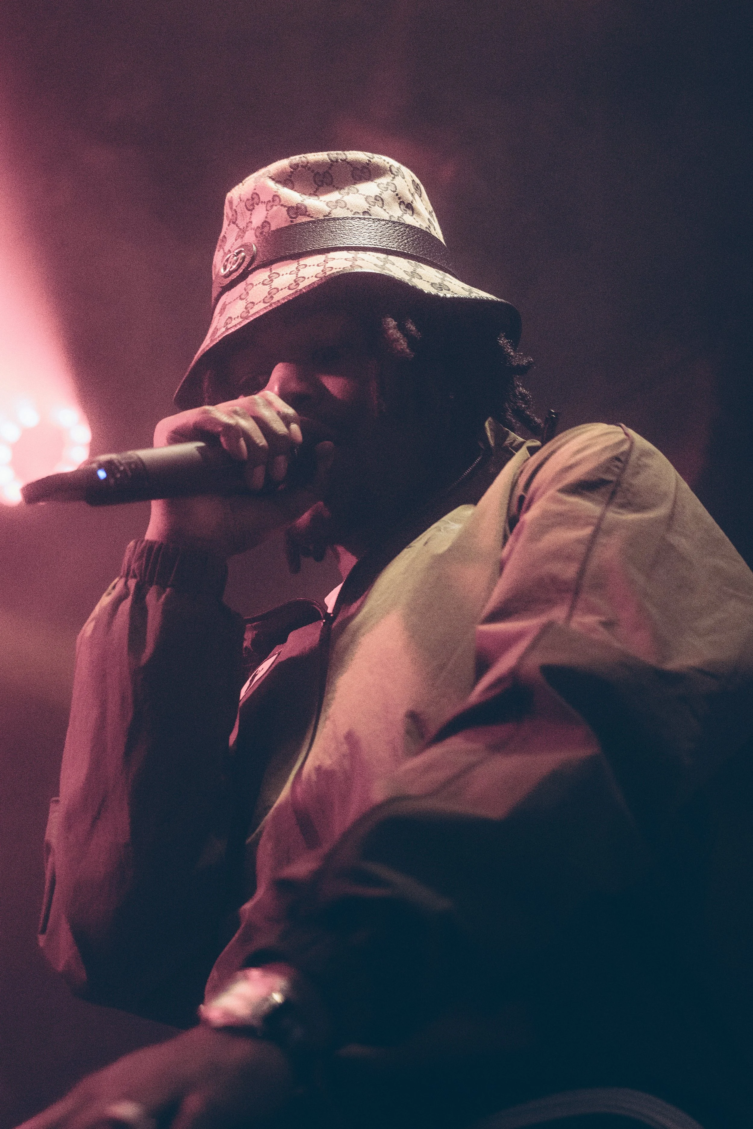 Un artiste de rap portant un chapeau Gucci, tenant un microphone lors d'une performance sous un éclairage rose et violet.